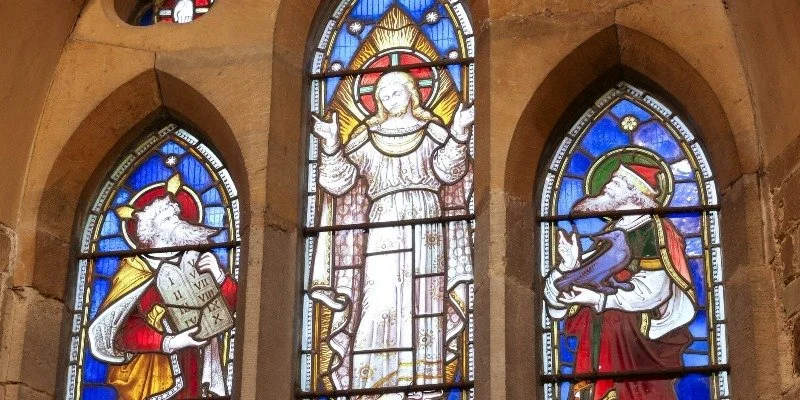 Three stained glass church windows depicting religious figures. The central window shows a woman dressed in white with a halo, arms raised, with sunlight behind her. The left window features a dog wearing a red cape or armor, holding a scroll or sword, with a halo. The right window shows a fox dressed in a military uniform with a red cape, holding a scroll or book, also with a halo.