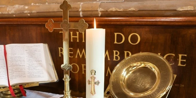 A Christian altar setup with a lit candle, a Bible, a silver cross, and a silver dish, against a wooden background with decorated letters spelling 'HIDE DO REMA BBCE'.