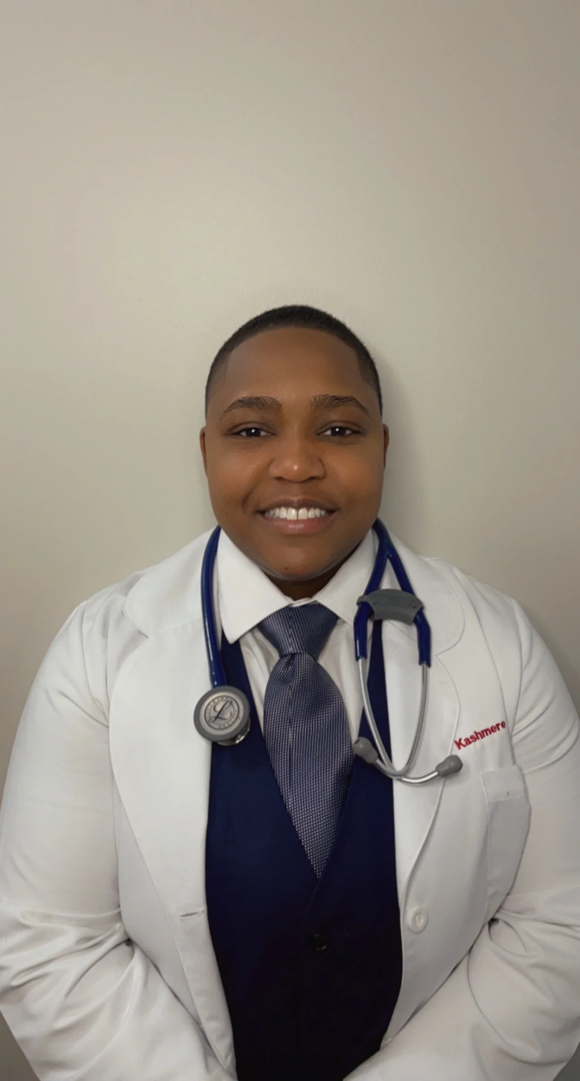 A young person in medical attire smiling, wearing a white lab coat with a stethoscope around their neck and a navy blue tie. The name tag on the coat reads 'Kashmere'.