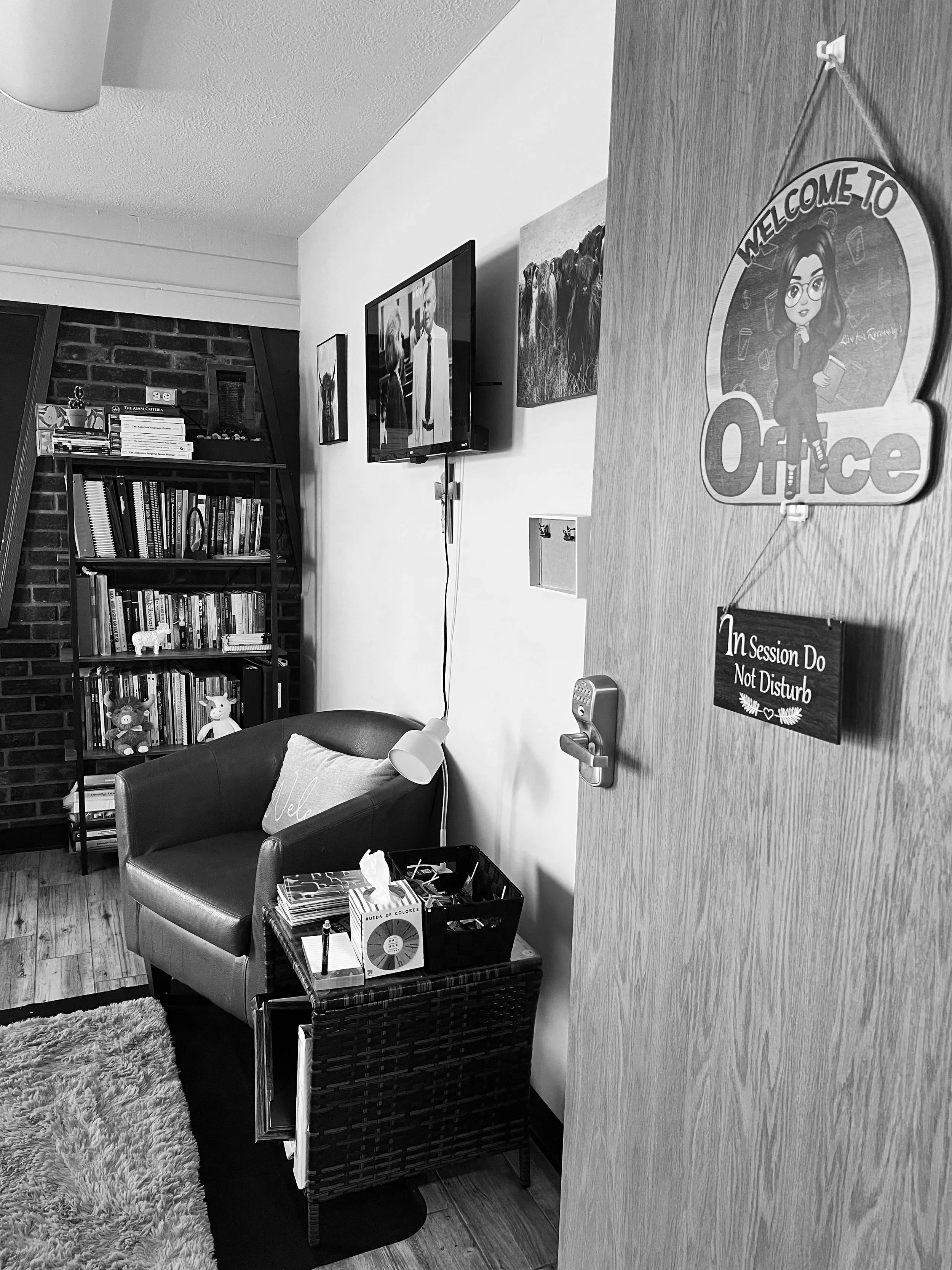 View of a cozy room with a bookshelf filled with books and toys, a small armchair with a cushion, a side table with magazines and office supplies, a wall-mounted TV, and a door with signs and decorations including a 'Welcome to Office' sign and a 'In Session Do Not Disturb' sign.