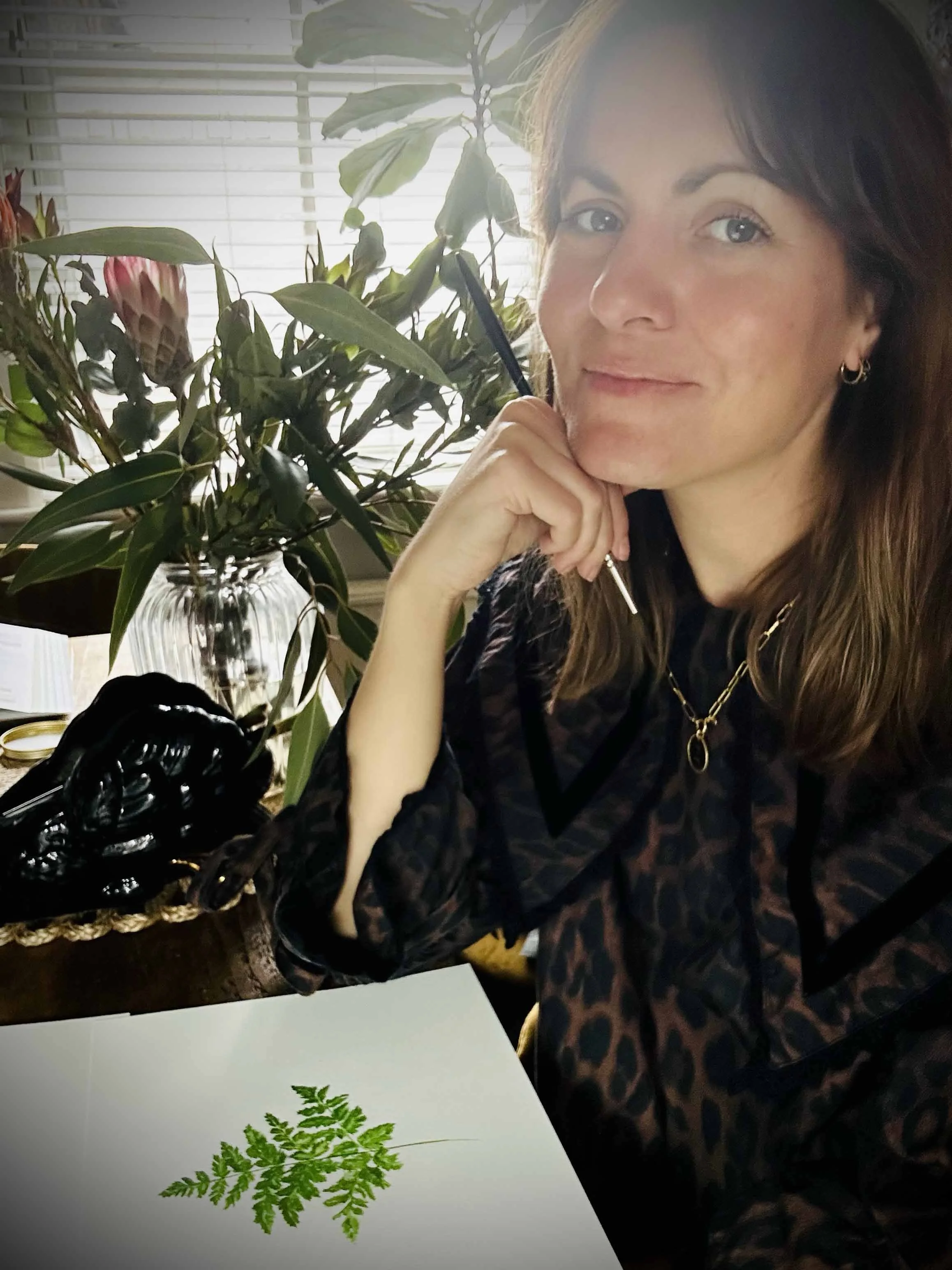 The artist sitting at a table who has brown hair and earrings holding a paintbrush. There are  a jar of flowers, and a paper with a green fern leaf print,  near a large plant in a windowed room.
