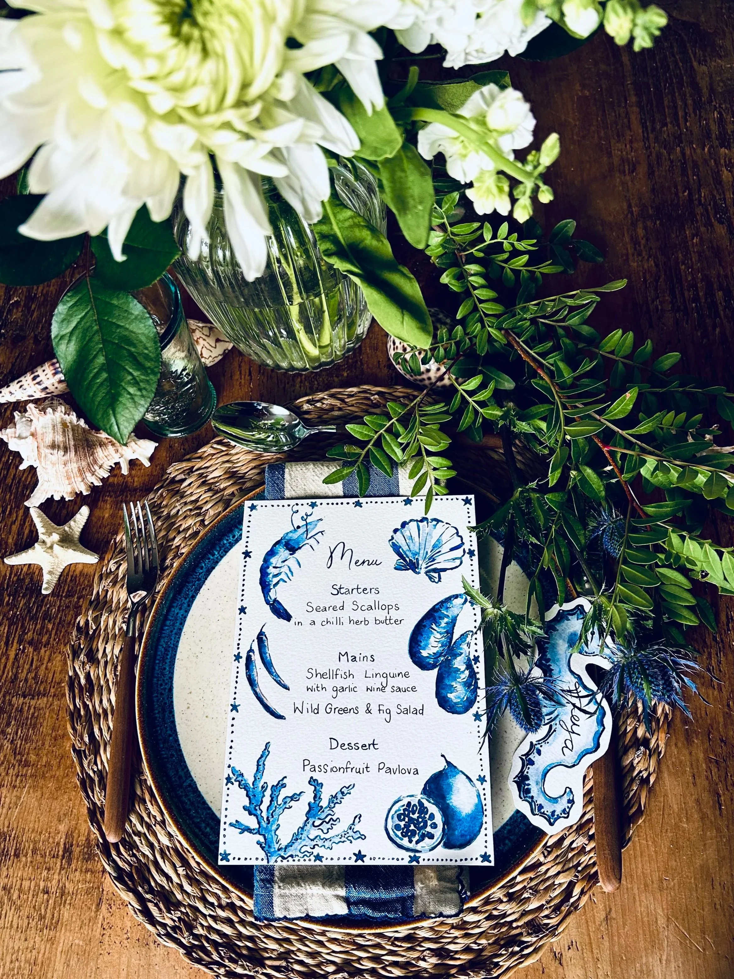 Table setting with a white plate, floral and vegetable decorations, a printed menu, a candle labeled 'Fresh Fig', and various green vases and foliage.