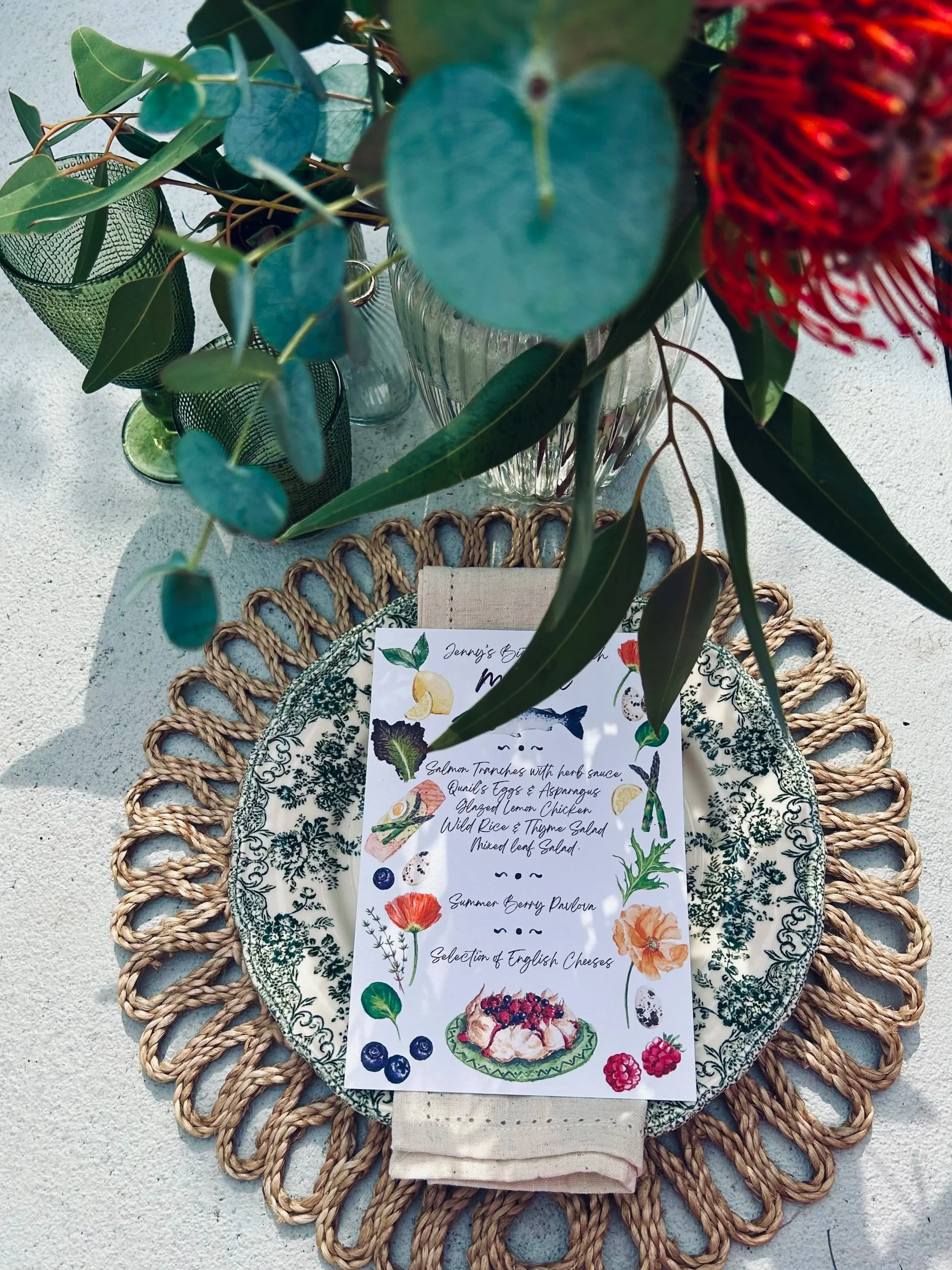 A place setting with a decorative plate, cloth napkin, and a menu card on top, surrounded by a woven placemat. A glass vase with green foliage and flowers is positioned above the setting.