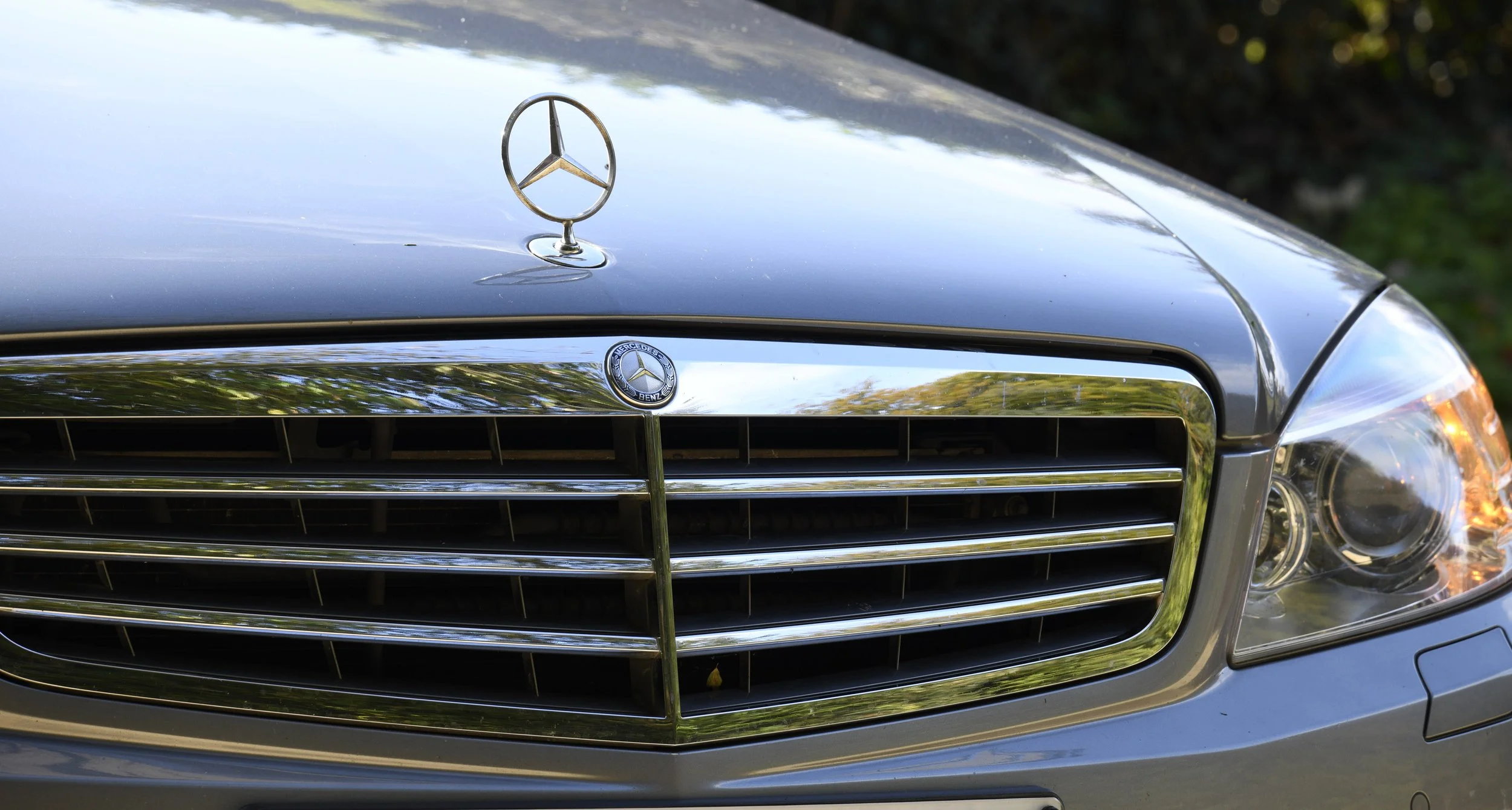Close-up of a black Mercedes-Benz car front, featuring the Mercedes-Benz emblem on the hood and grille, with part of the headlight visible.