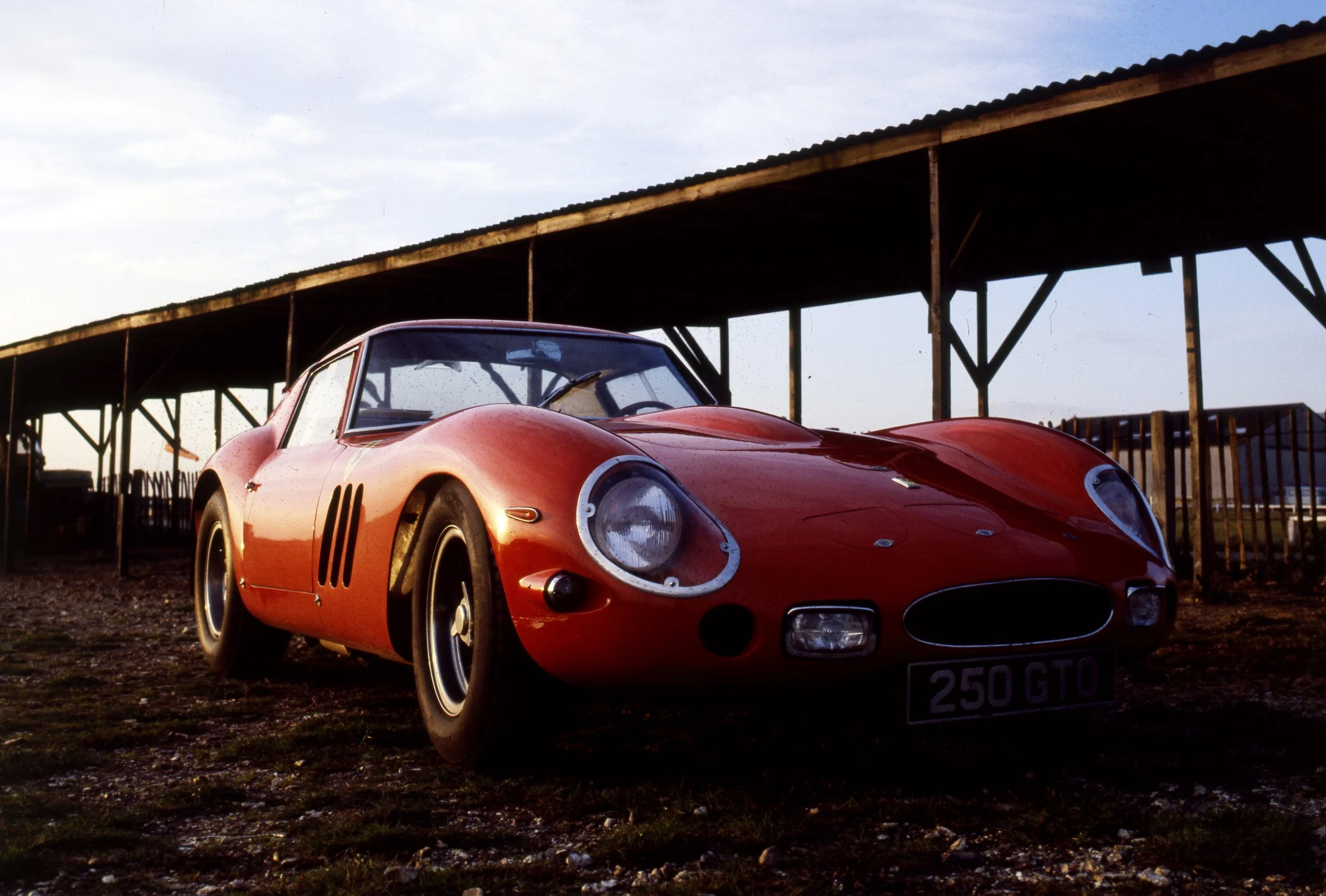 A red vintage race car parked outdoors under a shelter with a wooden roof, during sunset or dusk.
