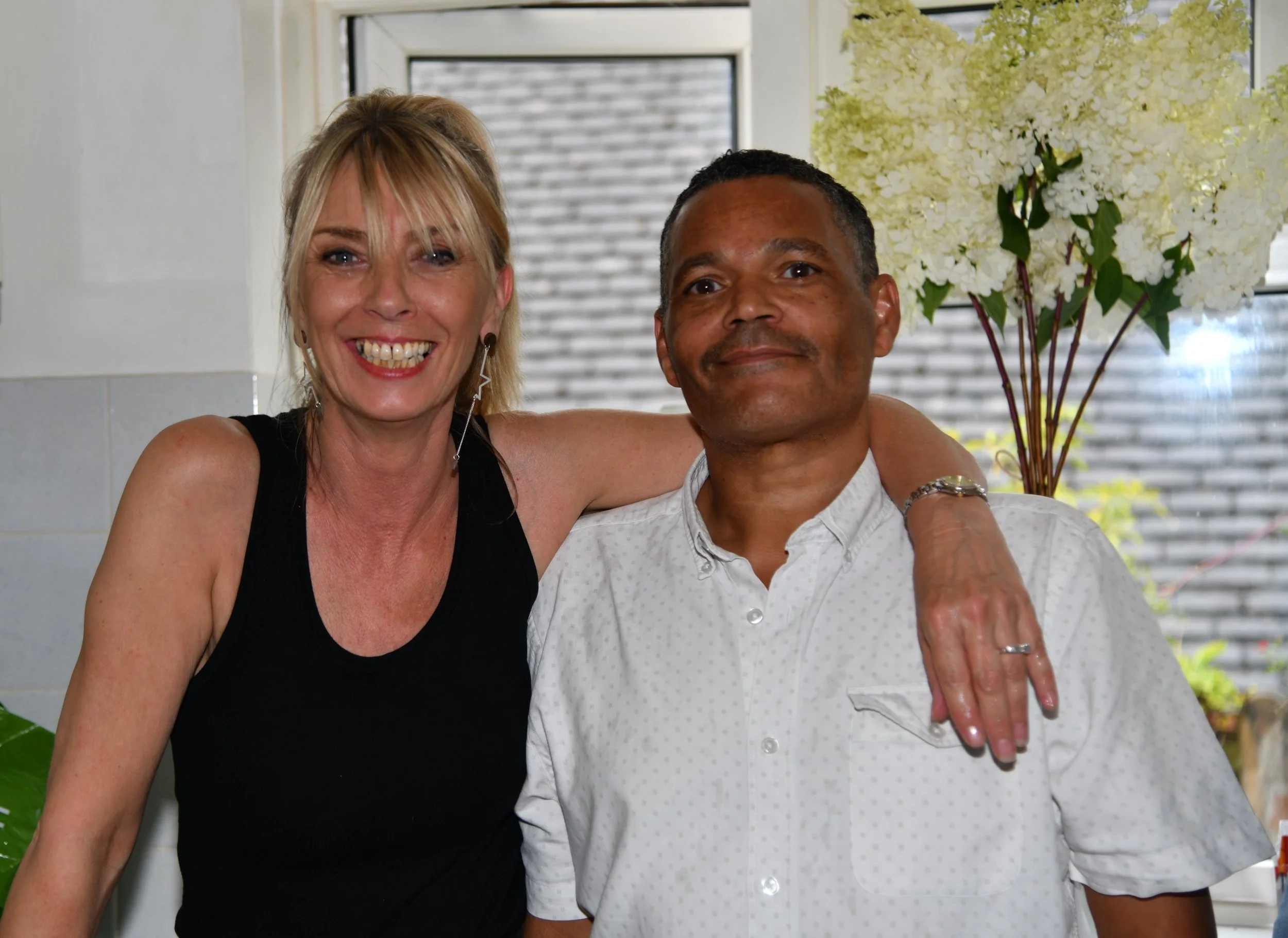 Two people smiling and posing for a photo indoors. The woman on the left has blonde hair, wearing a black sleeveless top, earrings, and is smiling widely. The man on the right has dark hair, wearing a white button-up shirt, with an arm around the wom