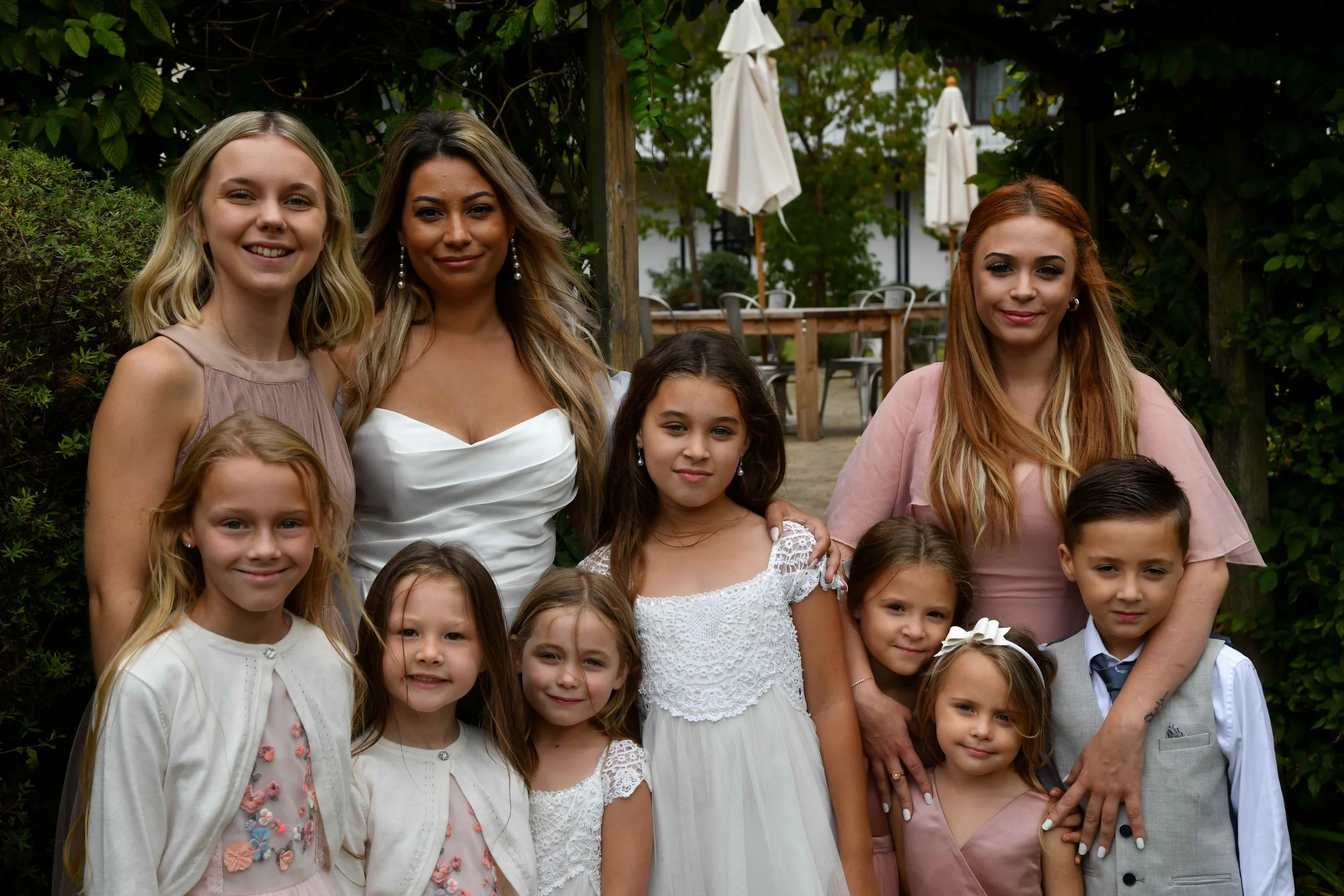 A group of women and children posing outdoors, surrounded by greenery and white umbrellas.