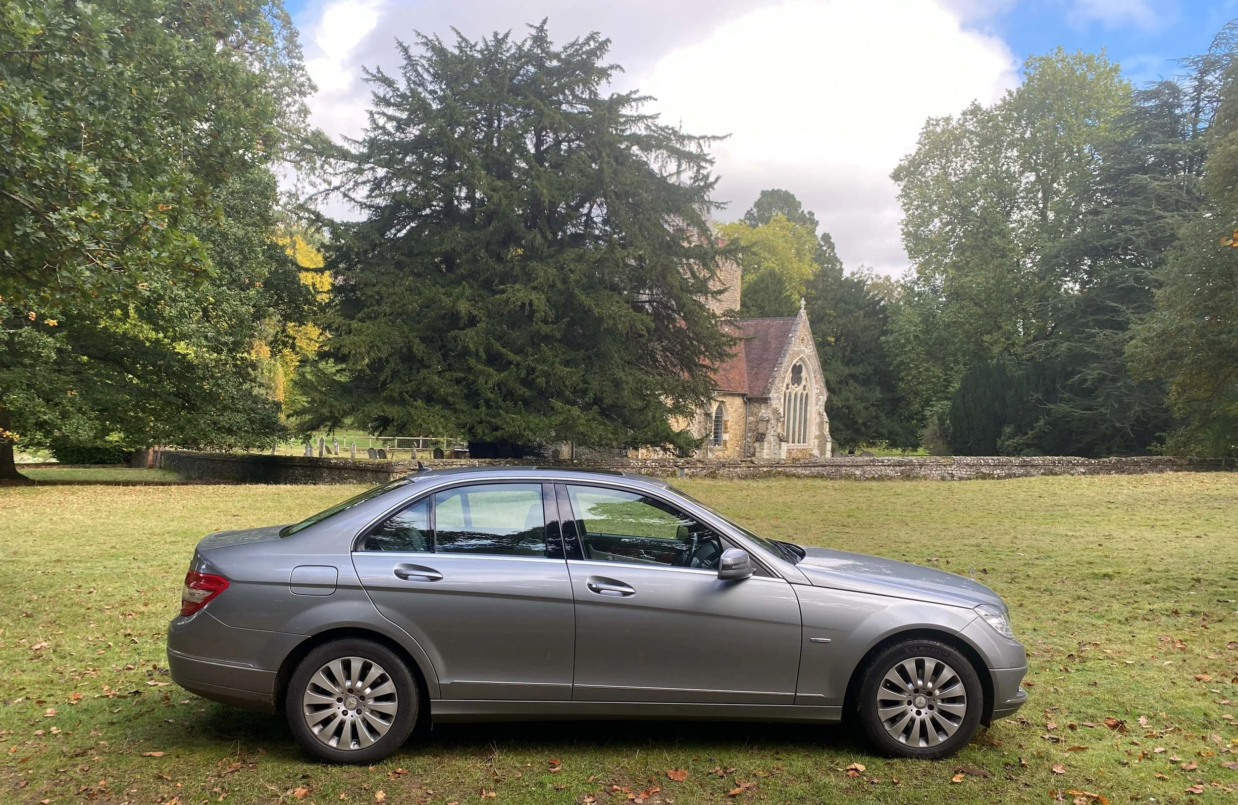 A gray sedan parked on a grassy area in front of a stone church with large trees surrounding it under a partly cloudy sky.
