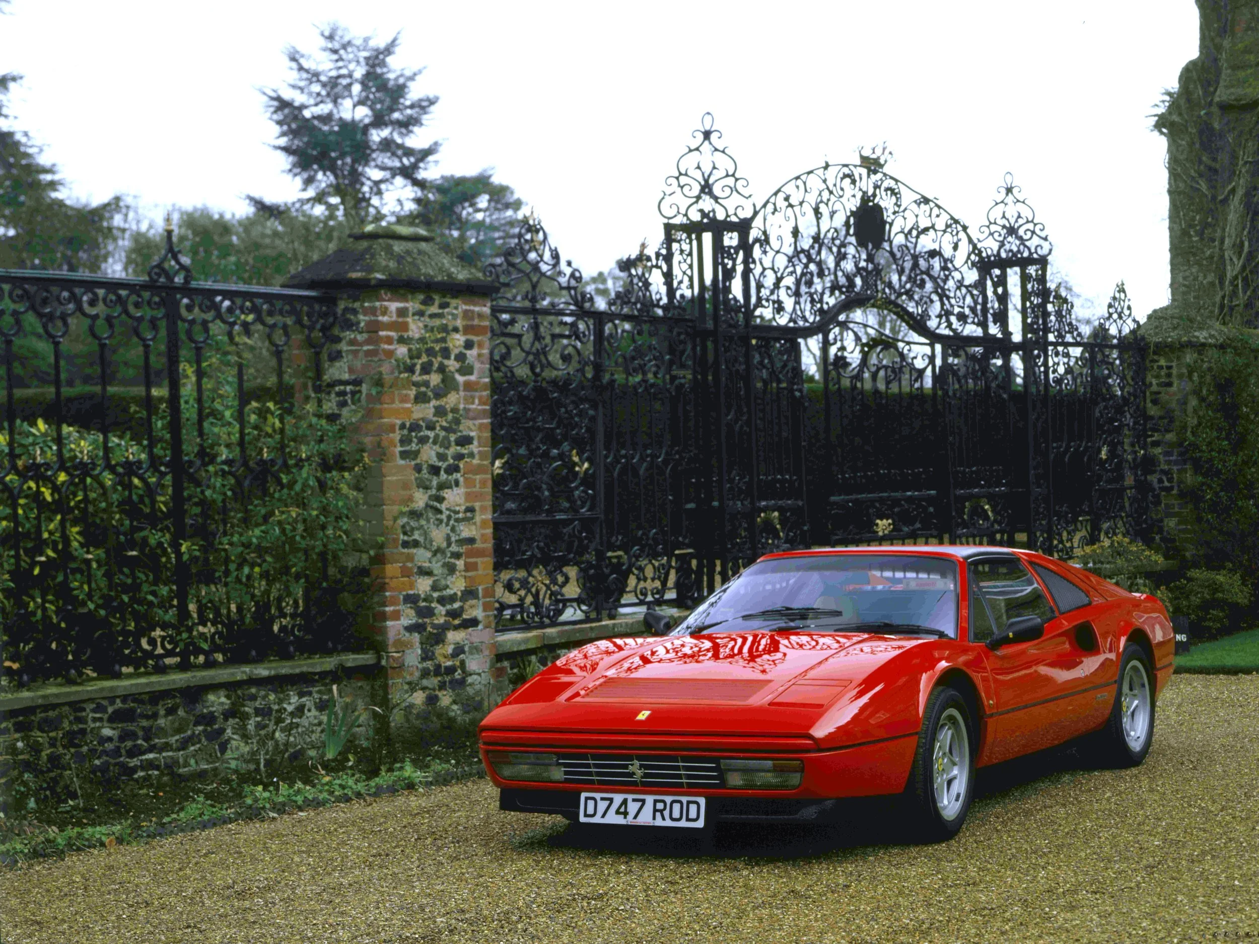 A red Ferrari sports car parked on a gravel driveway in front of an ornate black wrought iron gate with brick and stone pillars, with trees and shrubs in the background.