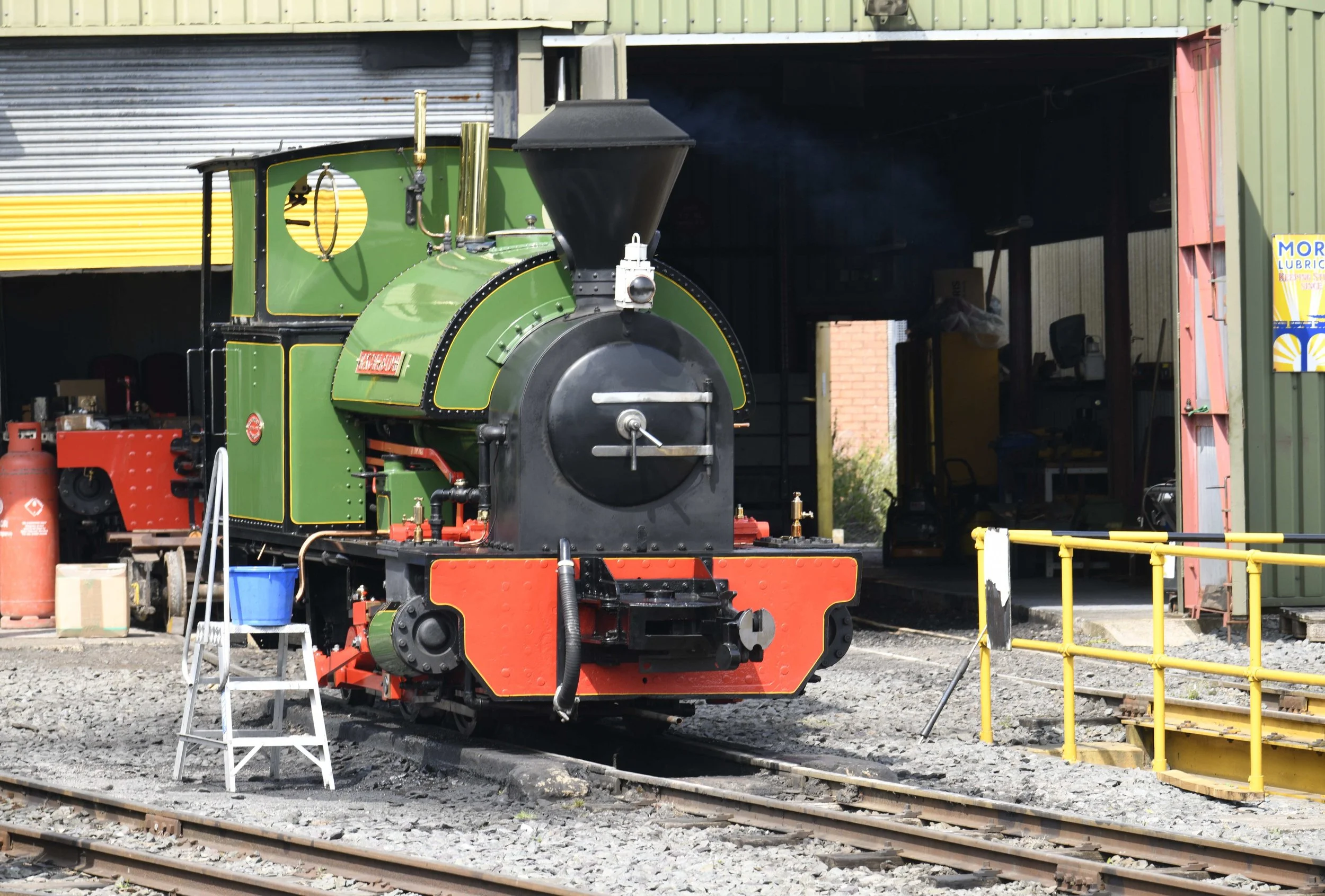 A vintage green steam locomotive with black and red details, positioned on railway tracks outside a workshop or storage shed with metal siding. There are tools and equipment nearby, including a step ladder, pipes, and a propane tank.
