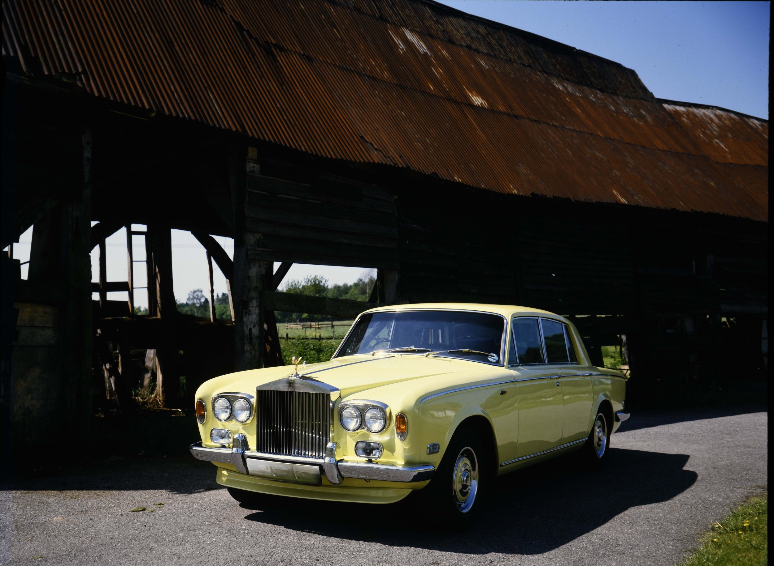A bright yellow vintage luxury car parked outdoors in front of a rustic barn with a tin roof and wooden walls.