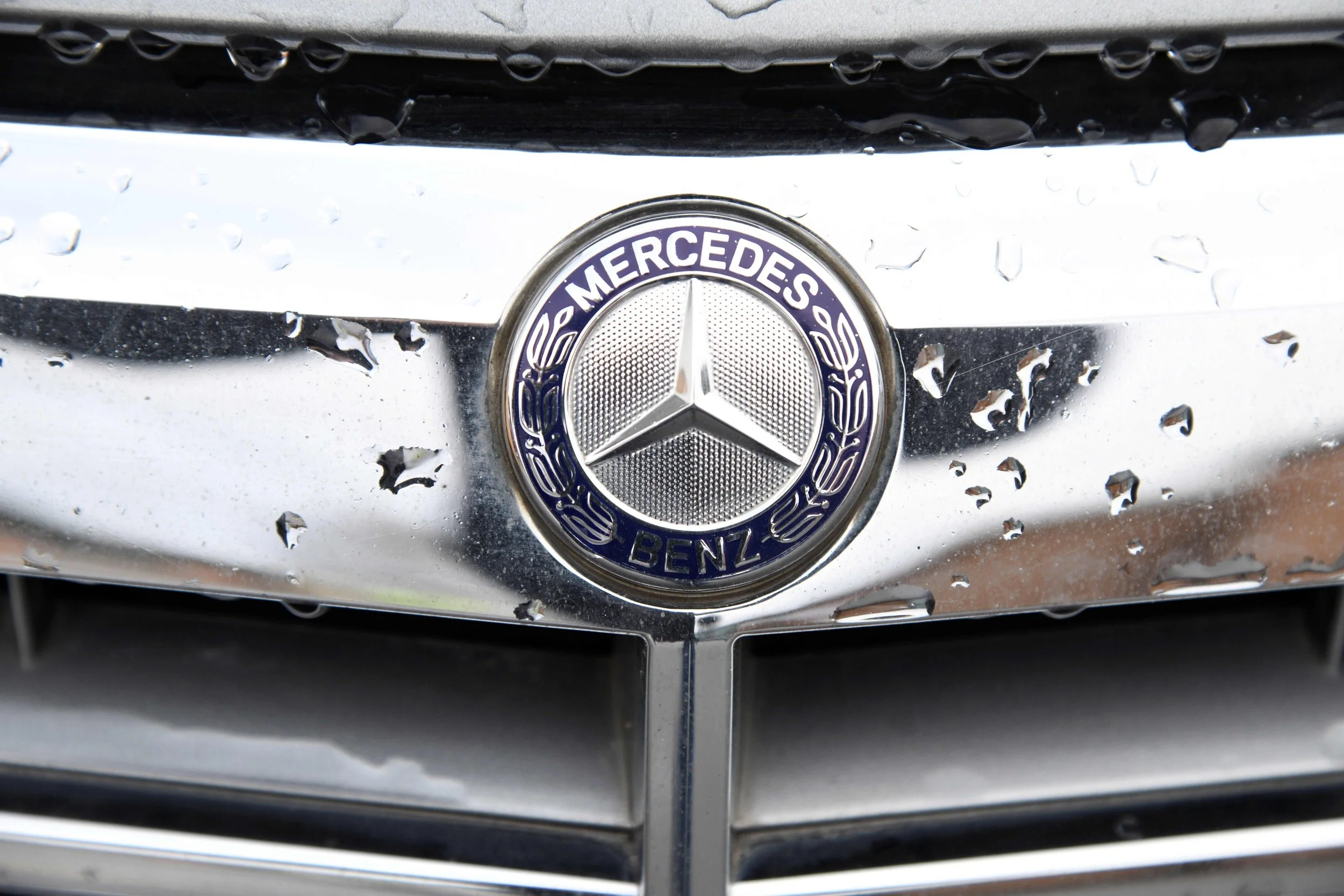 Close-up of a Mercedes-Benz logo on a chrome car grille covered in water droplets.