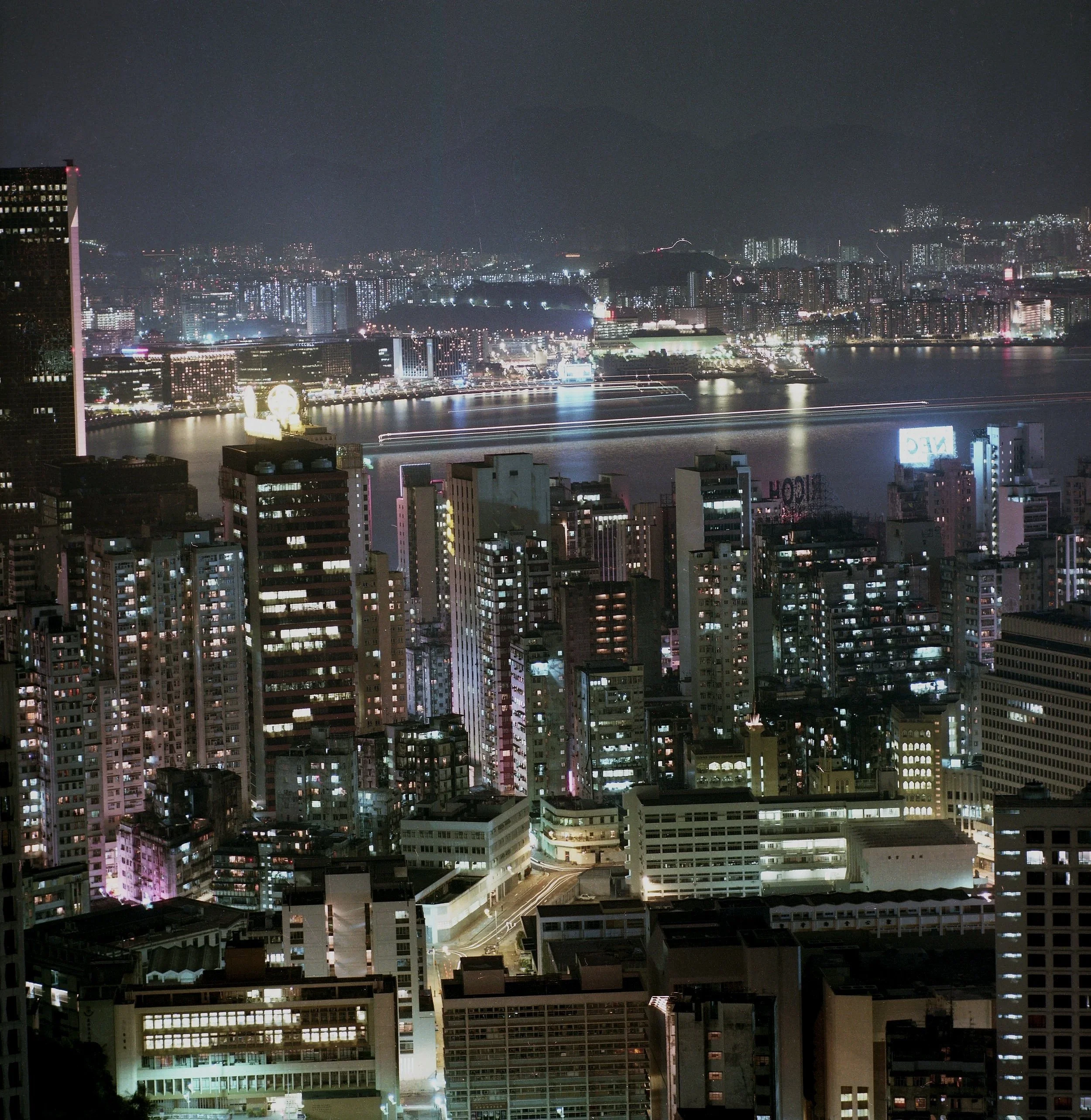 Nighttime cityscape with numerous high-rise buildings, illuminated windows, and a body of water reflecting city lights. Streets are visible with light trails from moving vehicles, and the background shows a dark sky.