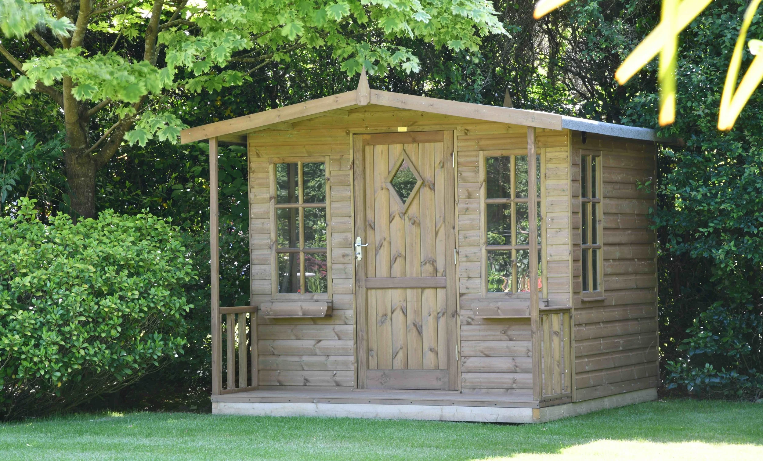 A small wooden garden shed beneath a lush leafy tree, surrounded by greenery.