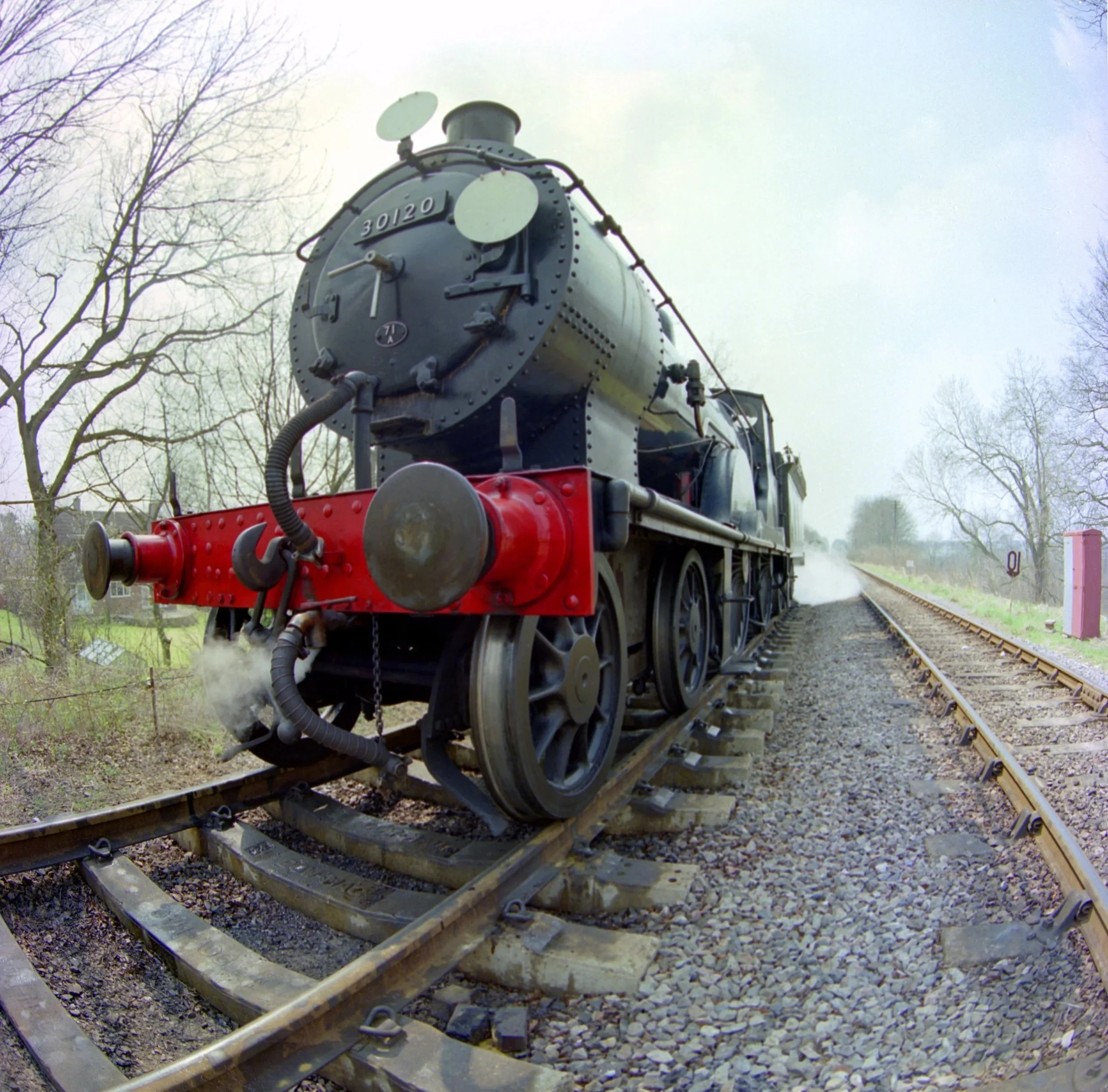 A steam locomotive on a railway track, viewed from a low angle, with bare trees and a cloudy sky in the background.