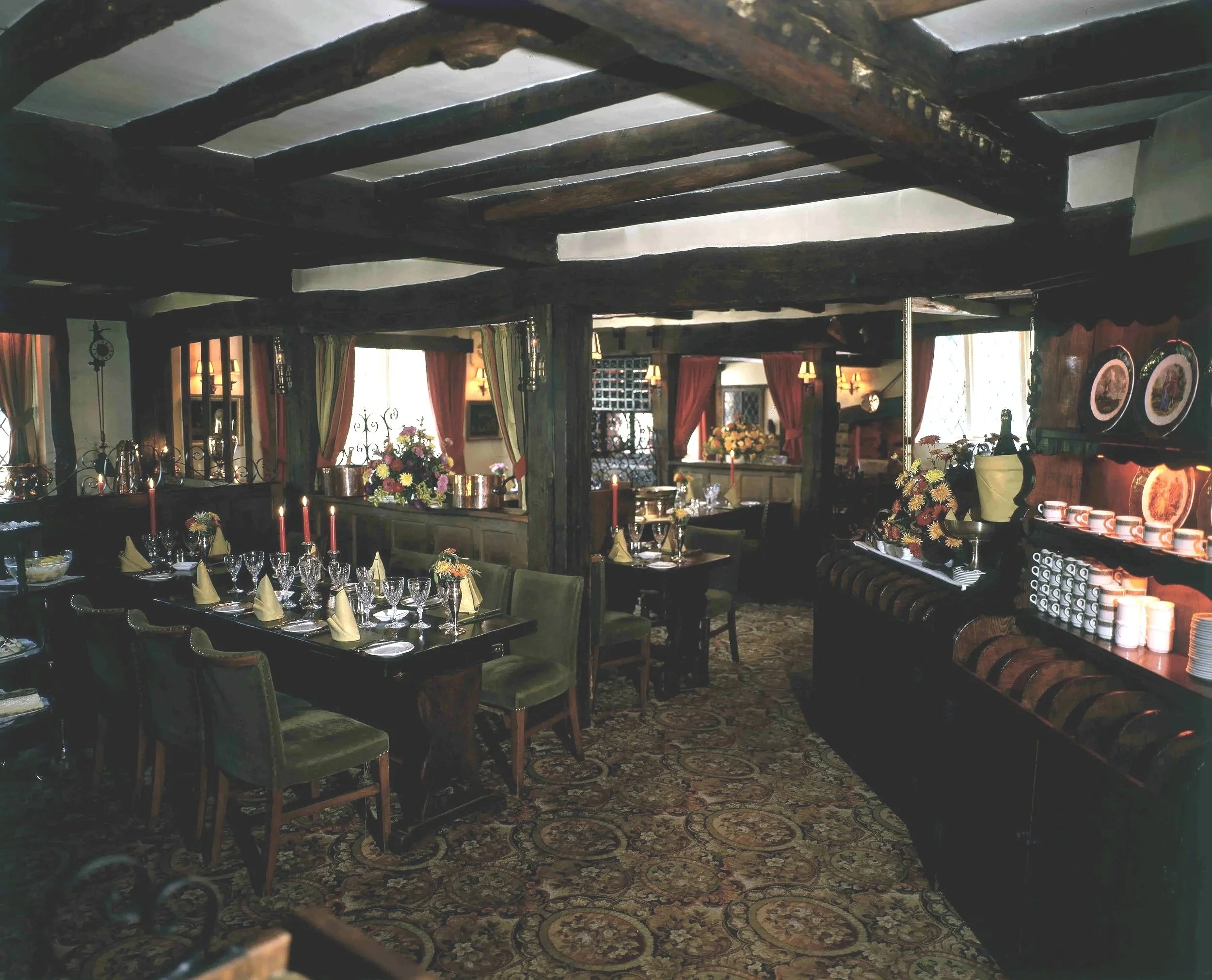 Interior of a rustic dining room with wooden beams on the ceiling, decorated with flowers, candles, and framed artwork. The room features vintage-style furniture, draped curtains, and a patterned carpet.