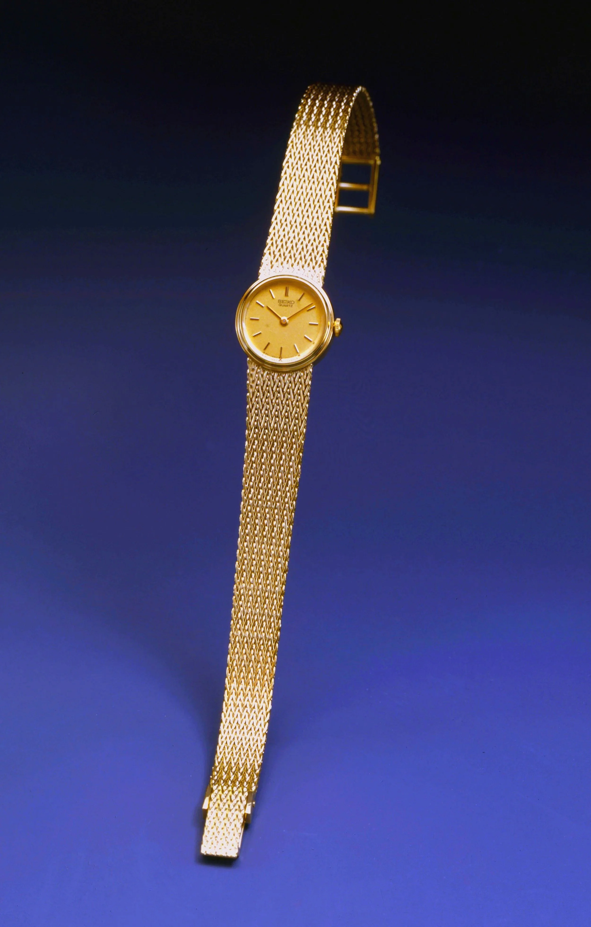 A gold wristwatch with a mesh band and a round gold face displayed on a blue background.