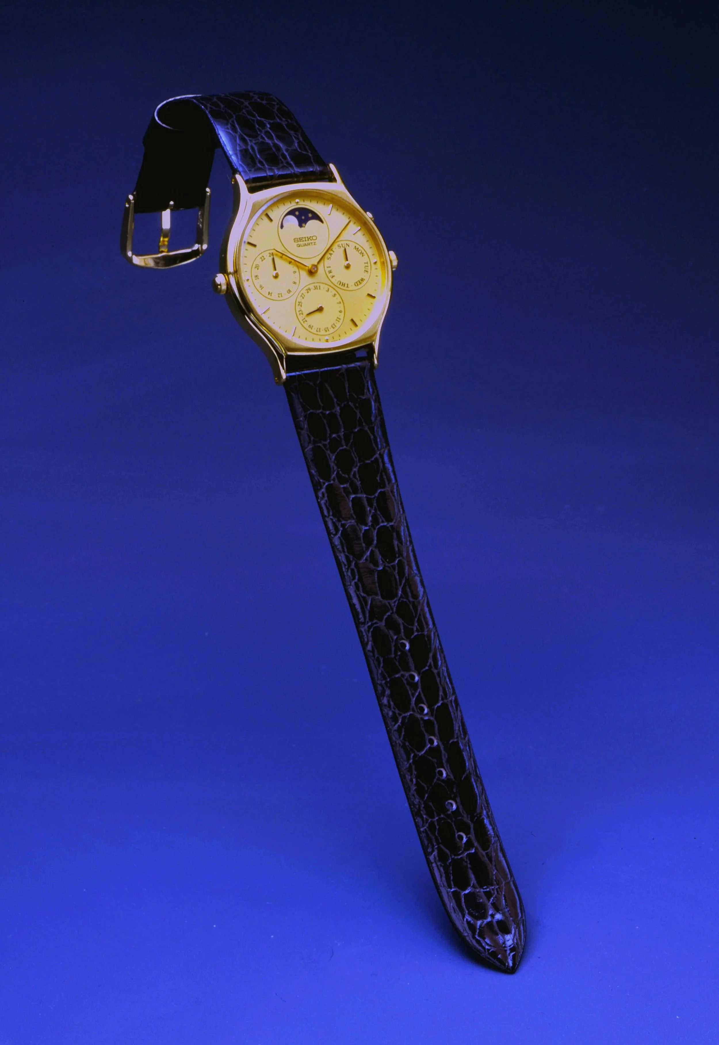 A luxury wristwatch with a black leather strap, gold case, and multiple dials on a dark blue background.