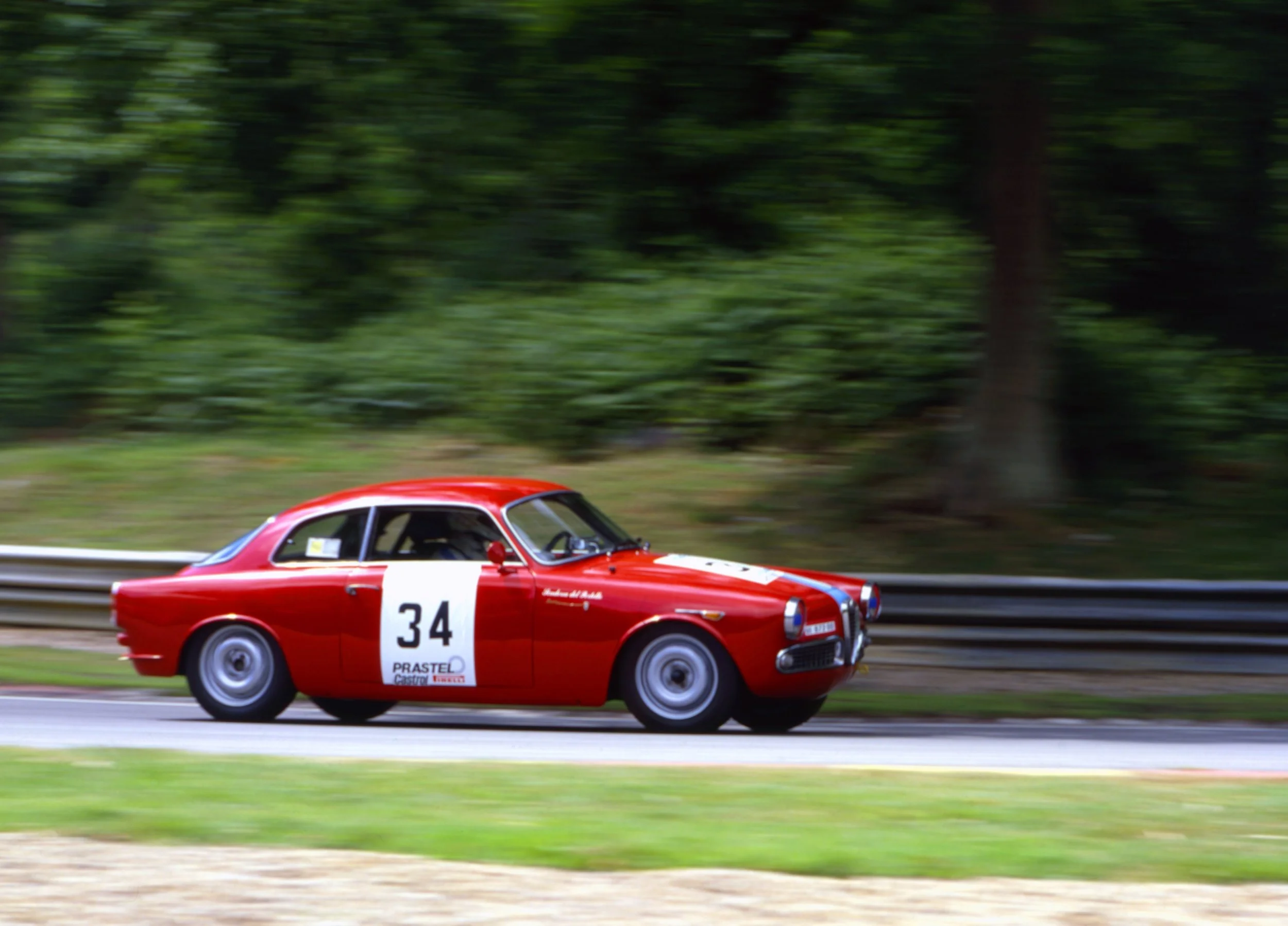 Red vintage race car with the number 34 on the side, racing on a track with green trees in the background.