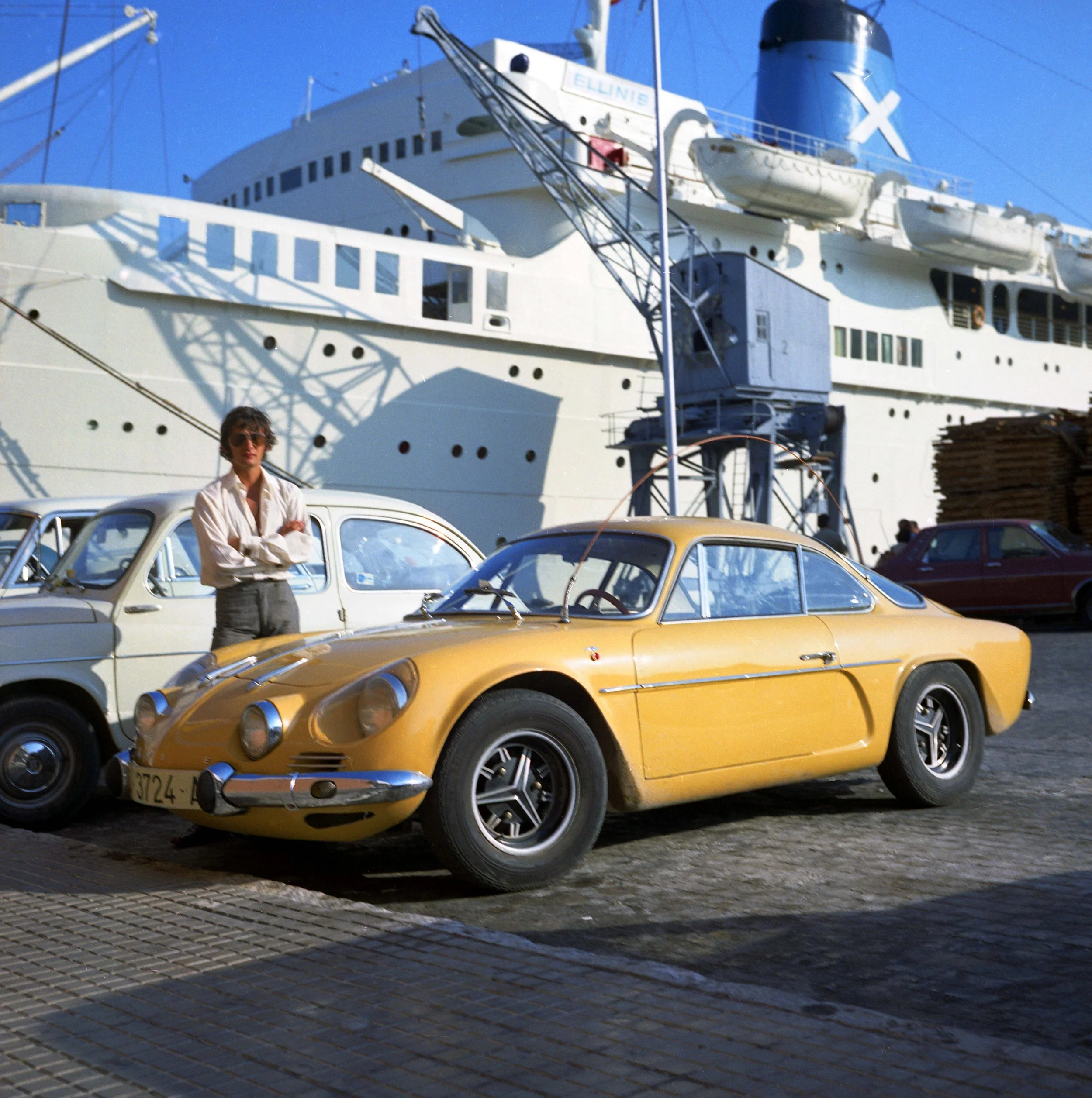 A woman with short hair and sunglasses standing in front of a row of vintage cars, with a large cruise ship docked in the background on a clear day.