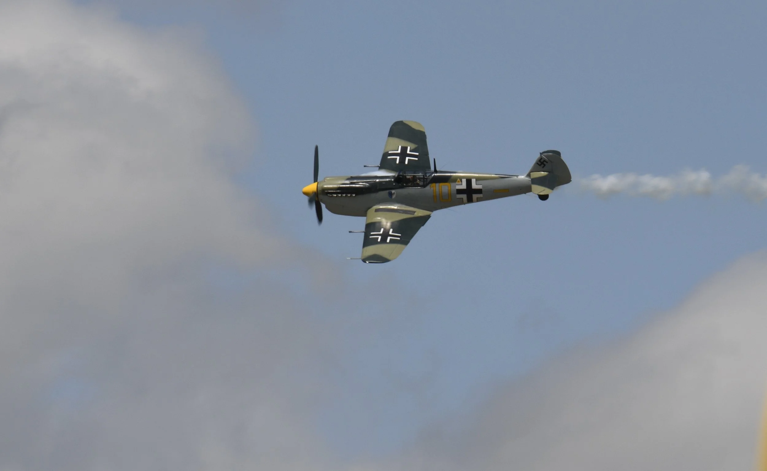 A vintage WWII-era fighter plane with black and white crosses on its wings and fuselage flying through a partly cloudy sky.