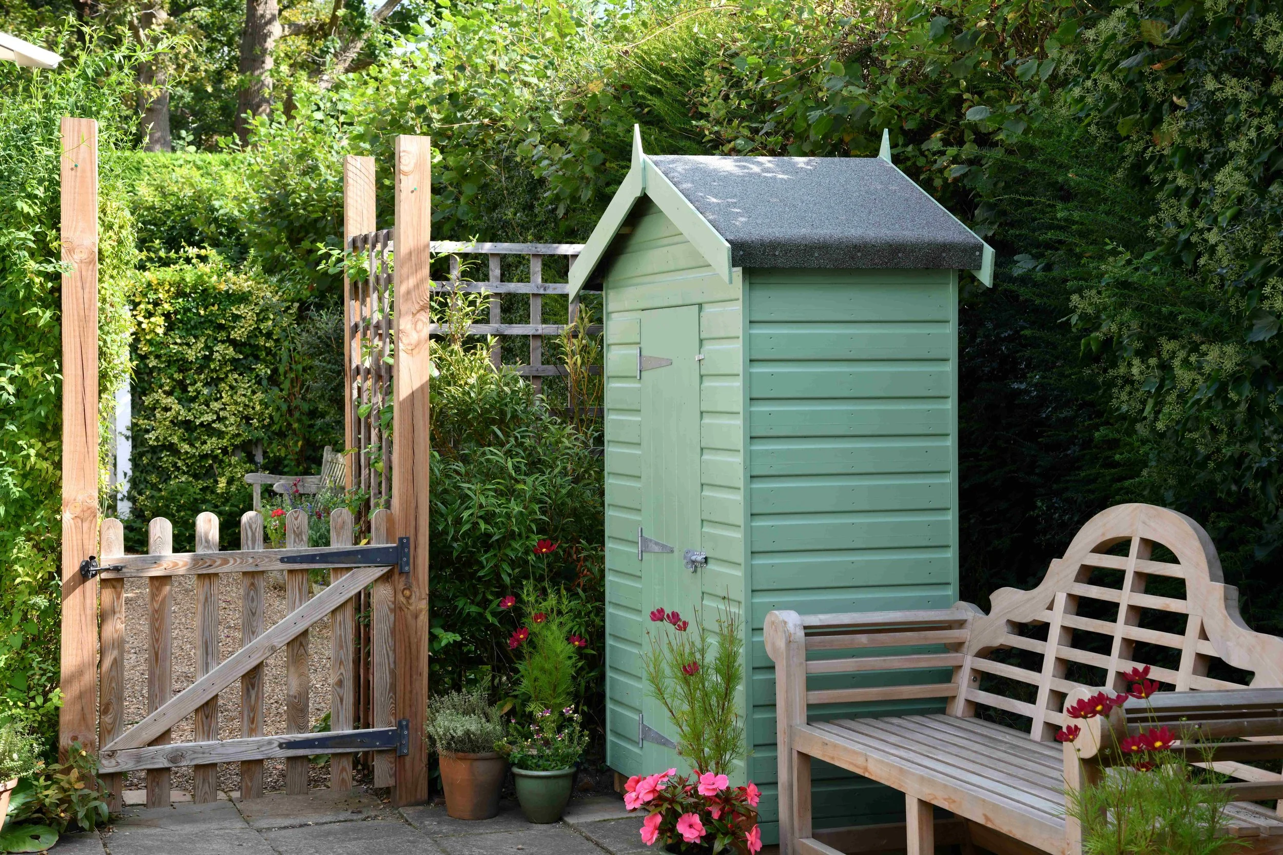 A backyard garden scene with a light green wooden shed, a wooden garden bench, flowering plants including pink and red flowers, potted plants, a small gate, and a natural fence made of wooden posts and lattice, all surrounded by lush green cohesive f