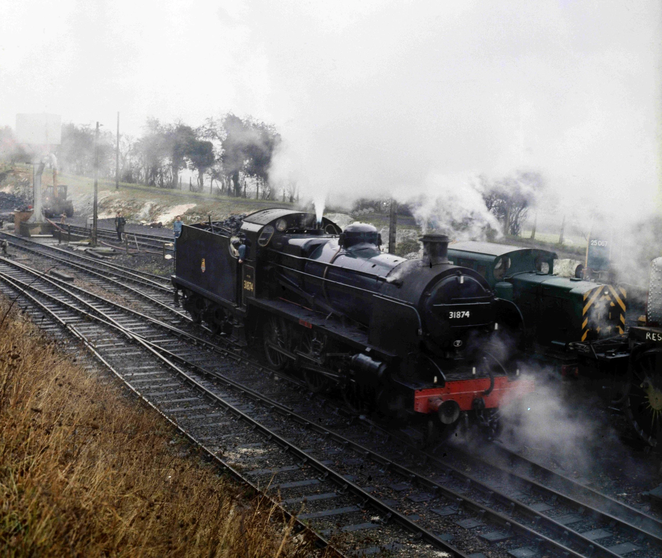 A steam locomotive on train tracks emitting black smoke, with a person walking nearby and trees and fog in the background.