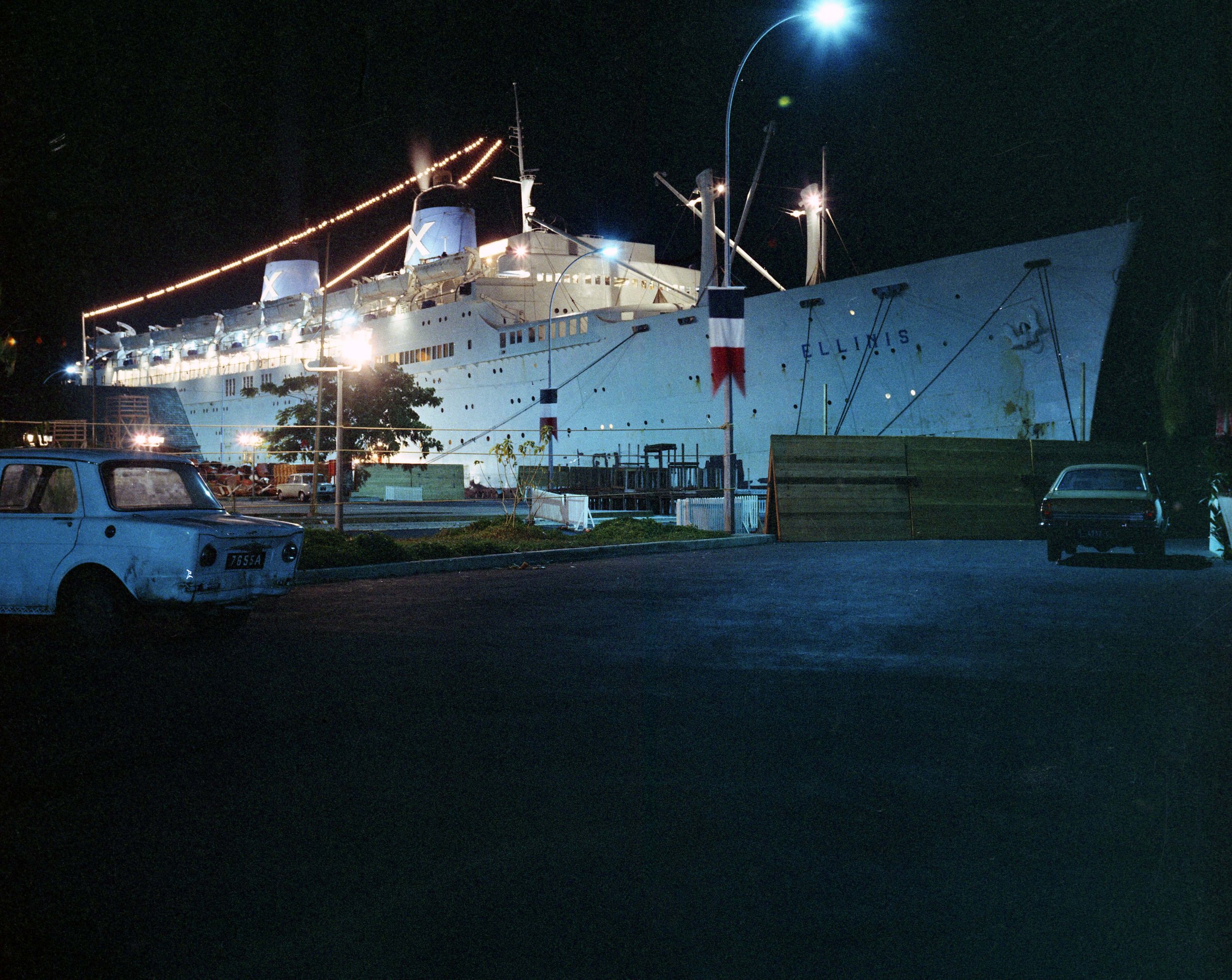 Nighttime view of a large white passenger ship named 'ELLIS' docked, with bright lights illuminating its structure. Several cars are parked in the foreground, and the sky is dark.