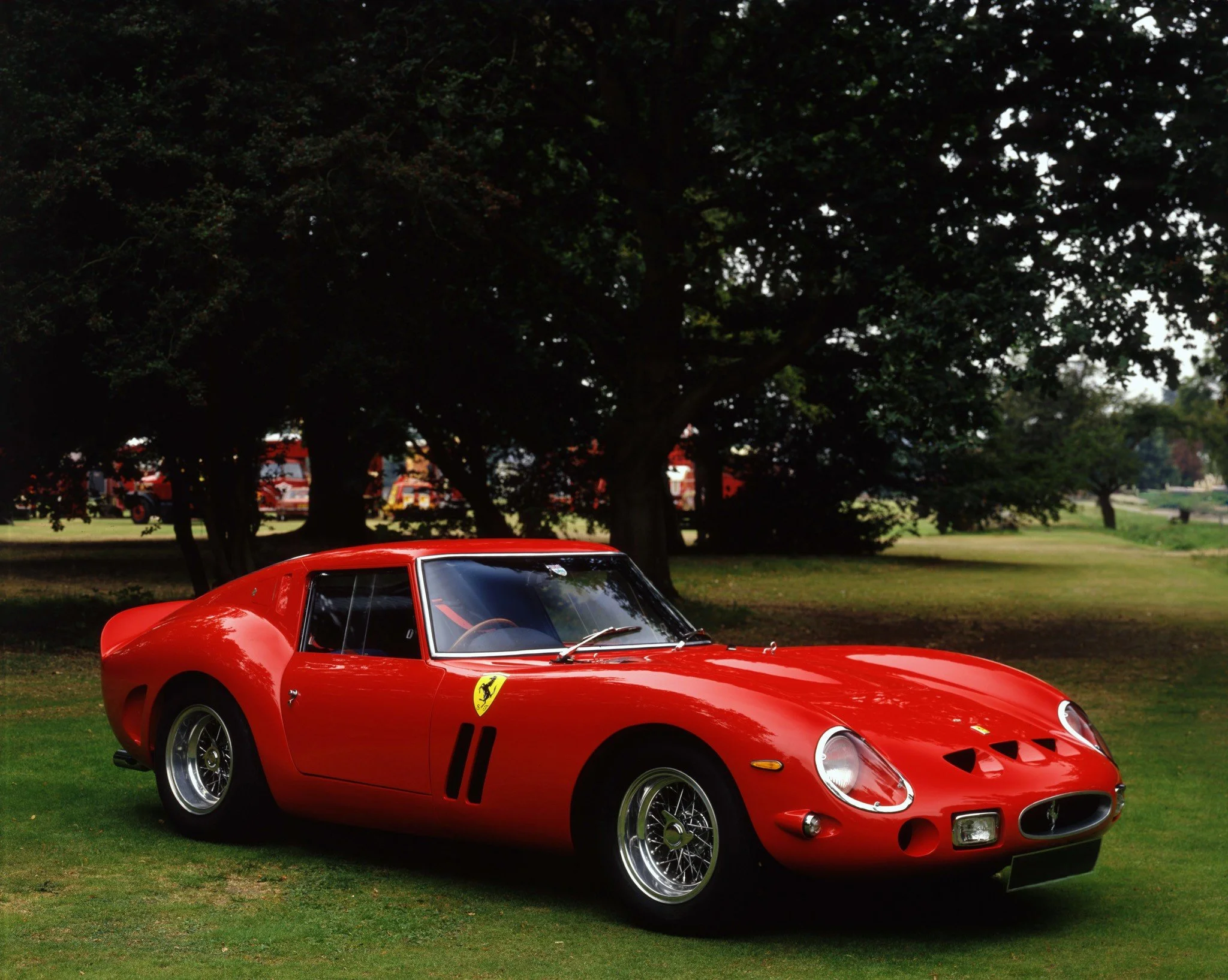 A red classic Ferrari sports car parked on a grass field with large trees in the background.