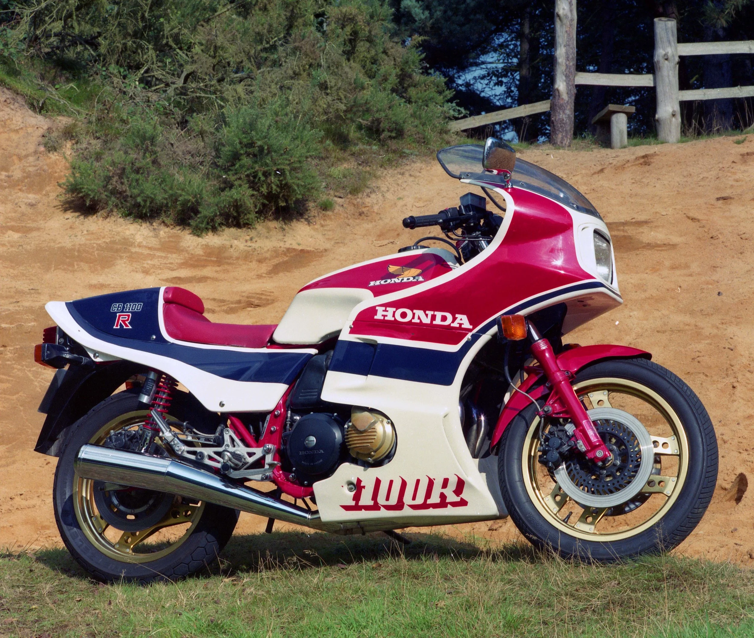 A vintage red, white, and blue Honda motorcycle with gold wheels parked on a grassy area with exposed dirt, surrounded by trees and a wooden fence in the background.