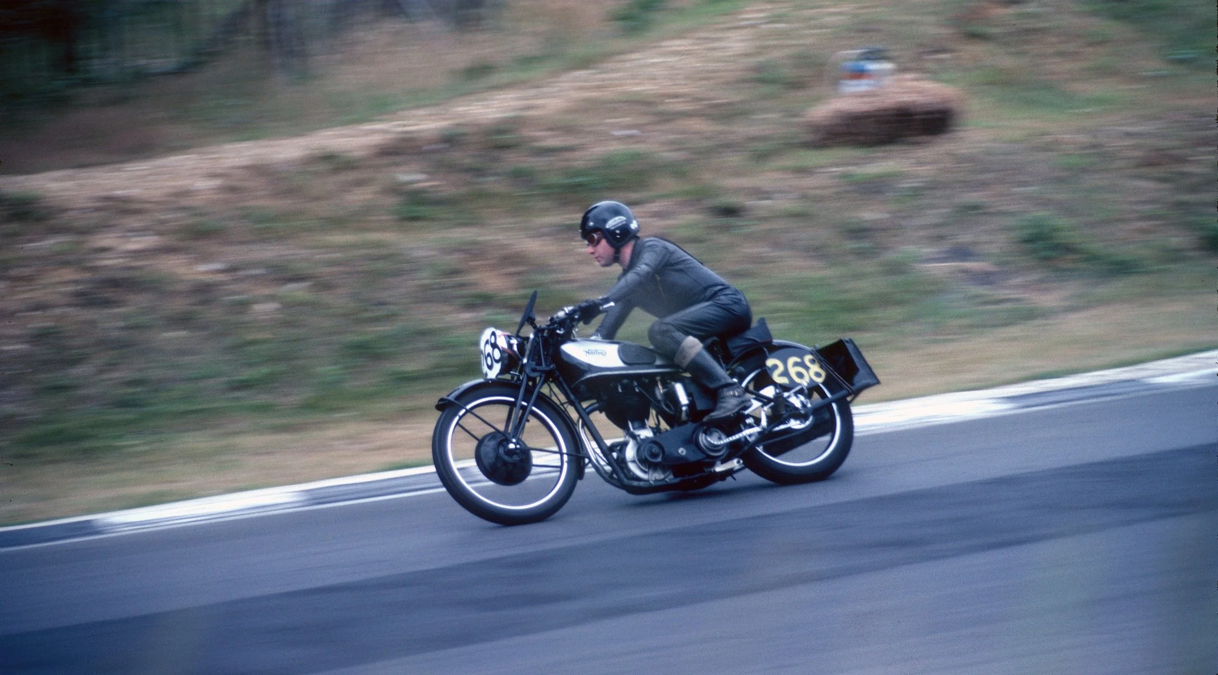 A person in black riding a vintage motorcycle with the number 268 on the side, wearing a helmet and sunglasses, on a paved road with a grassy hillside in the background.