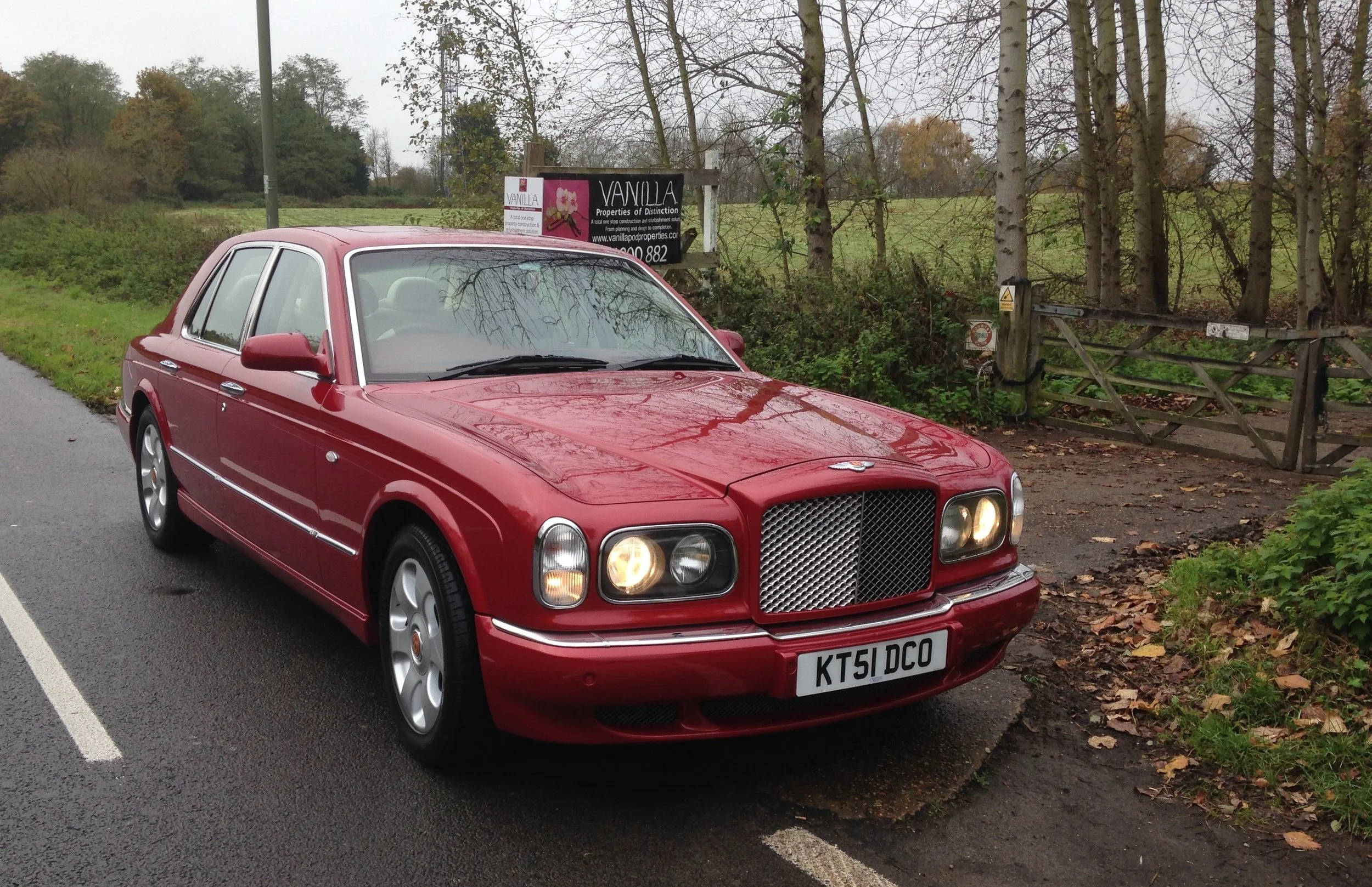 Red vintage Bentley car parked on the side of a wet road near a wooden gate and trees, with a sign advertising property development in the background.