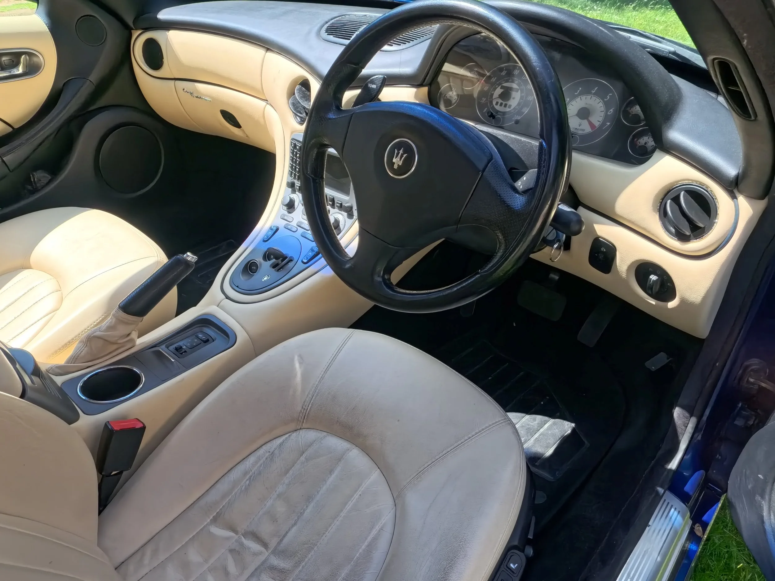 The image shows the interior of a car with cream-colored seats, a black steering wheel with a Maserati logo, and a dashboard with various gauges and controls. The car's interior is photographed from the driver's side, with sunlight visible on the sea