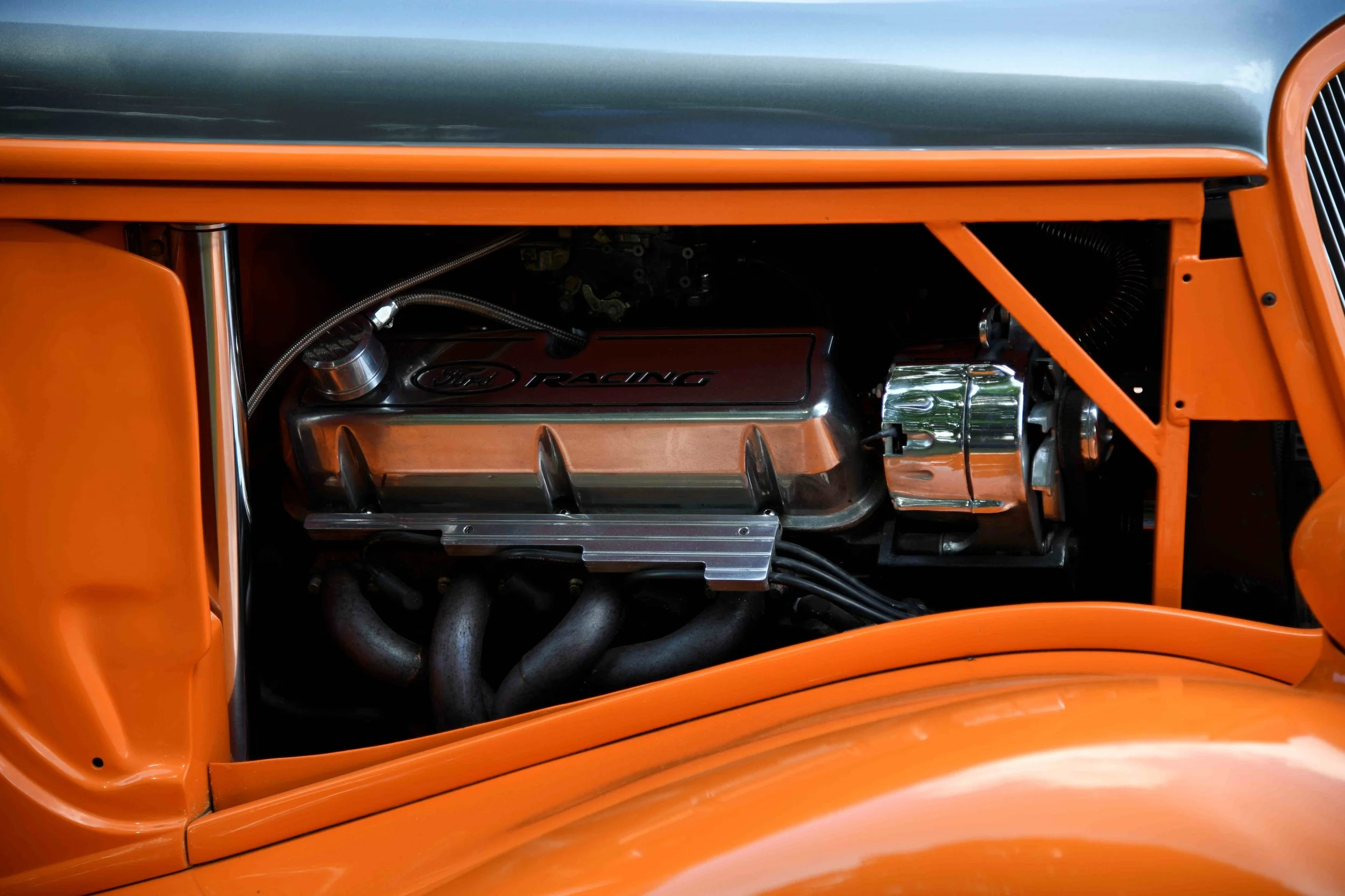 Close-up of a vintage race car engine with a silver hood and orange body, showing the engine cover marked 'Ford Racing'.