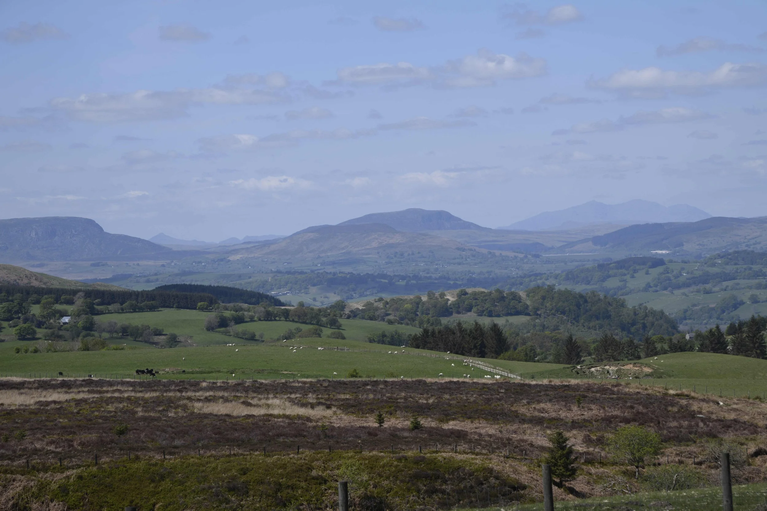 Scenic view of rolling green hills, scattered farmland, and distant mountains under a partly cloudy sky.