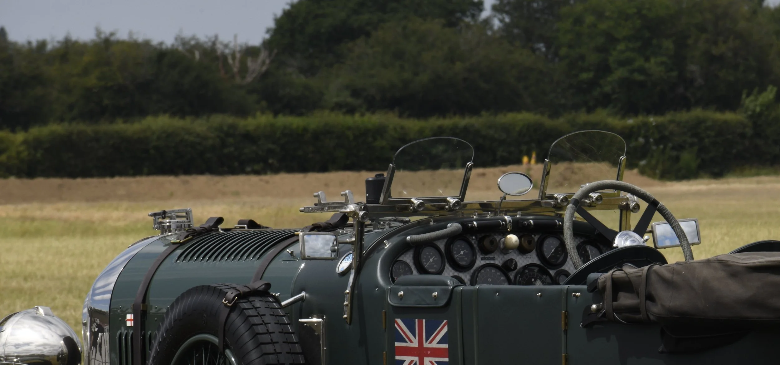 Close-up of a vintage racing car with a British flag sticker, multiple gauges, mirrors, and protective windshield, set outdoors on a grassy field with trees in the background.