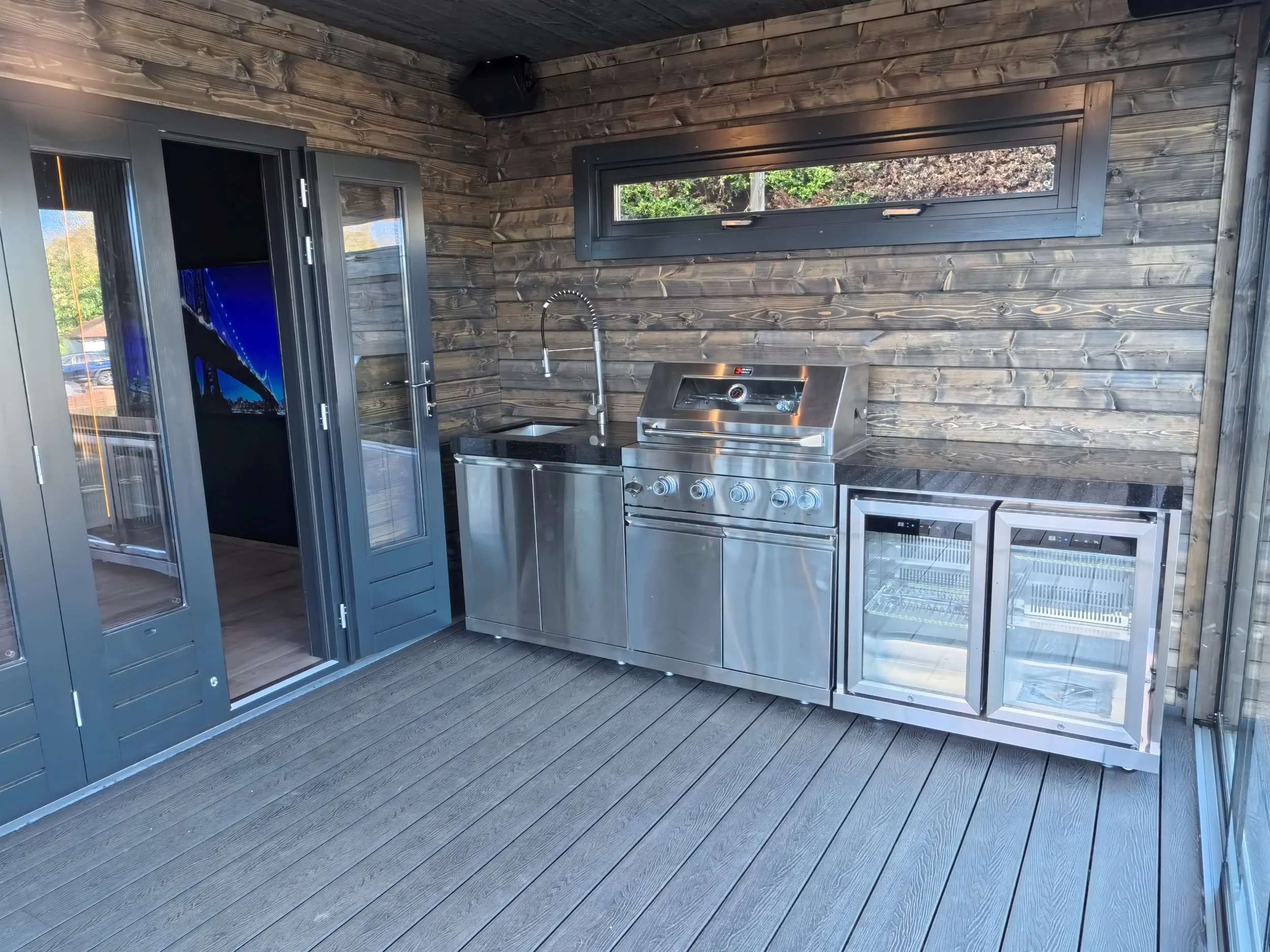 An outdoor kitchen with stainless steel grill, mini fridge, and sink, set against a wooden wall with a long horizontal window, on a wooden deck.