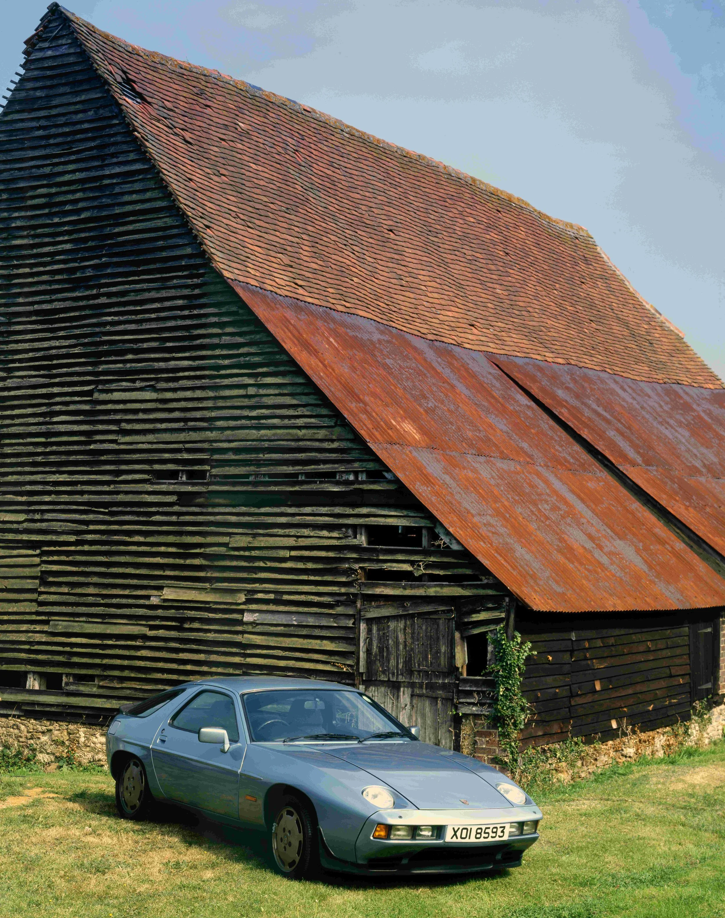 A silver vintage sports car parked on a grassy field in front of an old wooden barn with a weathered, rusty roof.