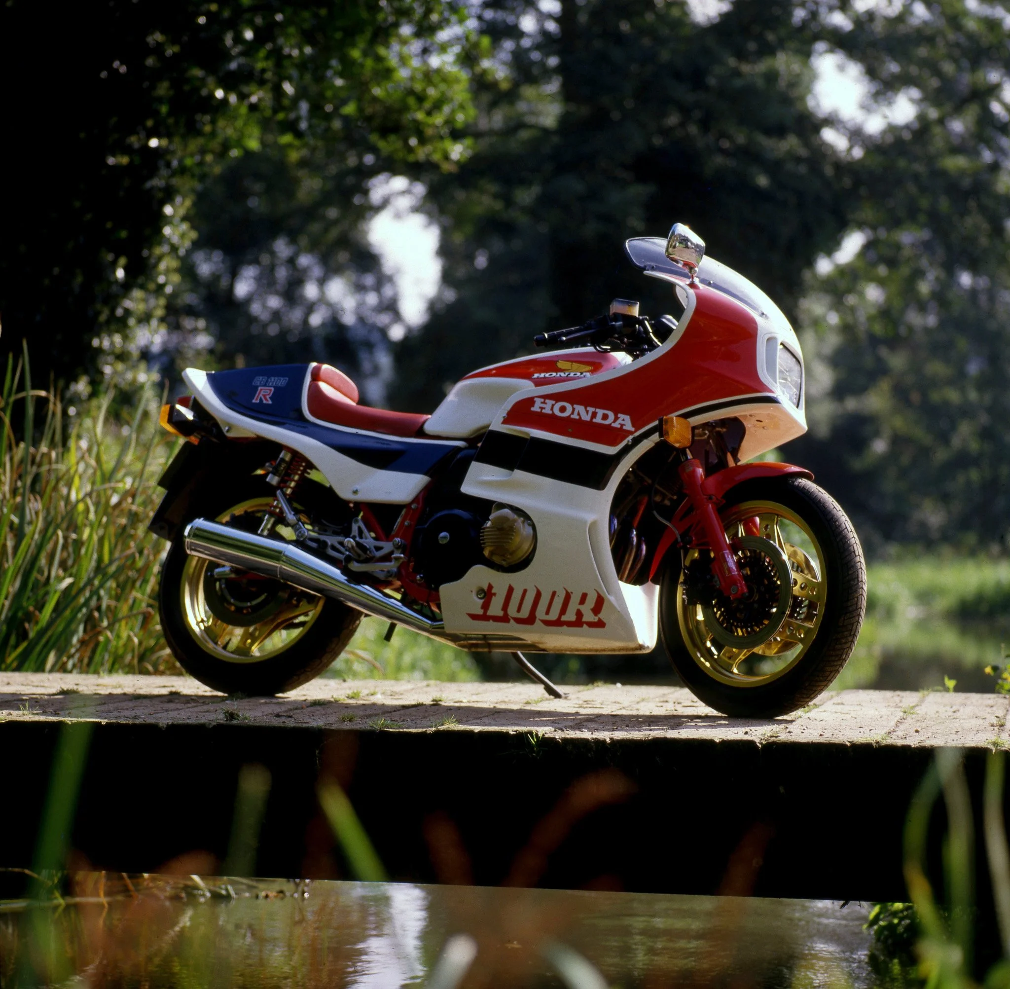 A red and white Honda motorcycle is parked on a path near a small creek with green grass and trees in the background.
