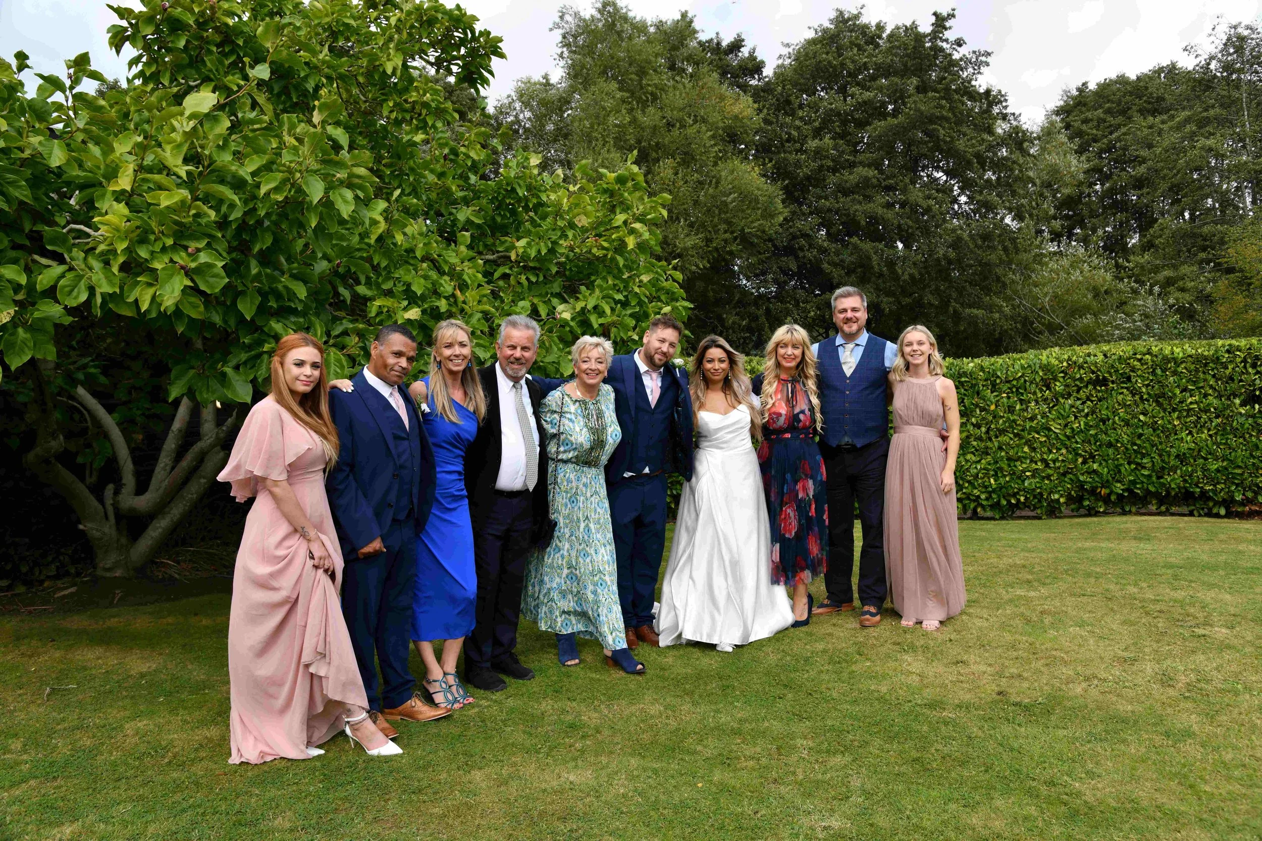Group of people dressed in formal attire standing outdoors on a grassy area with trees in the background.