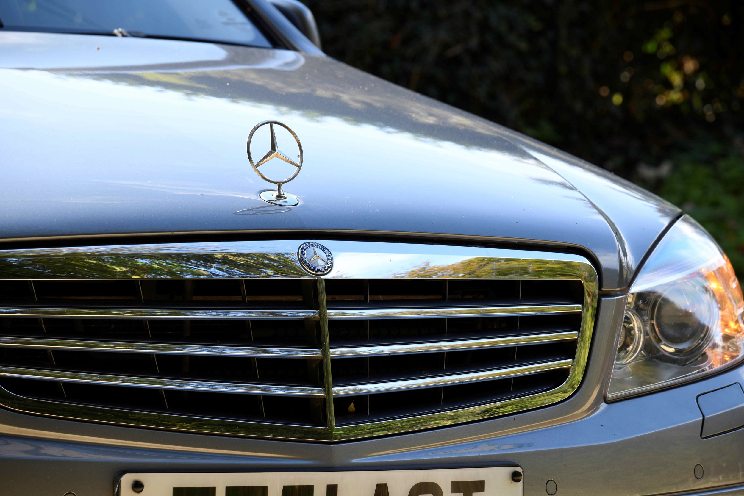 Close-up of a silver Mercedes-Benz vehicle hood with the Mercedes logo and grille, with a partially visible license plate.