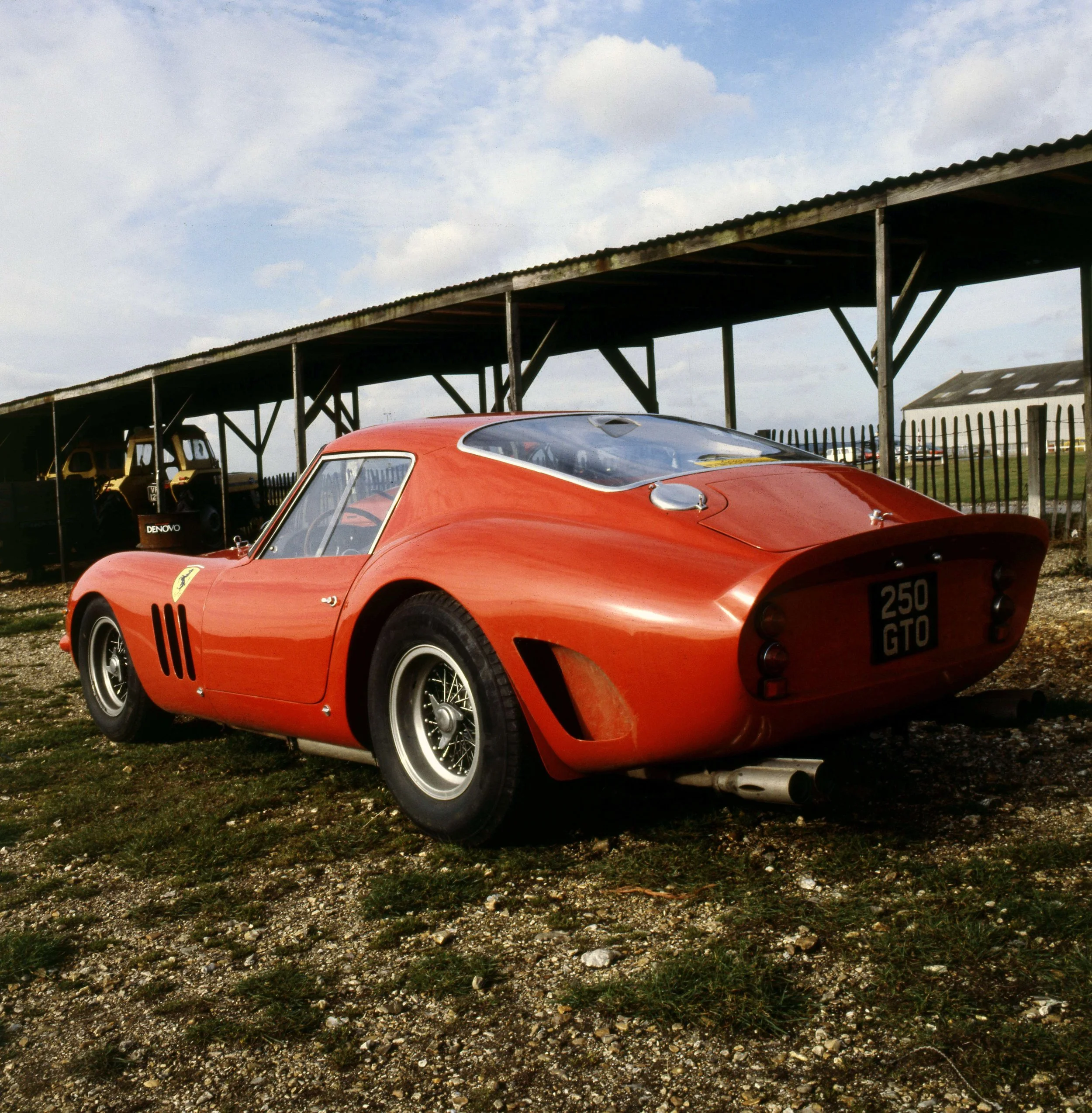 A red vintage racing car with a sleek design and a Ferrari logo on the side, parked outdoors on a grassy area with gravel. In the background, there is a metal shed or stable with a weathered roof, and some vintage vehicles and equipment. The sky is p