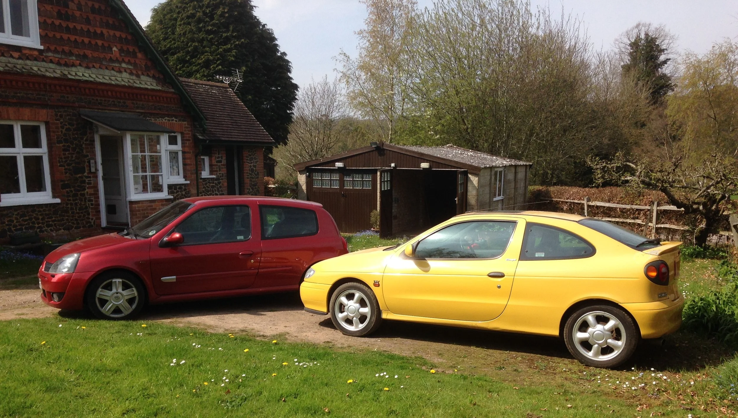 Two small cars, one red and one yellow, parked on a grassy area in front of a brick house with white-framed windows and a nearby wooden shed surrounded by trees and bushes.