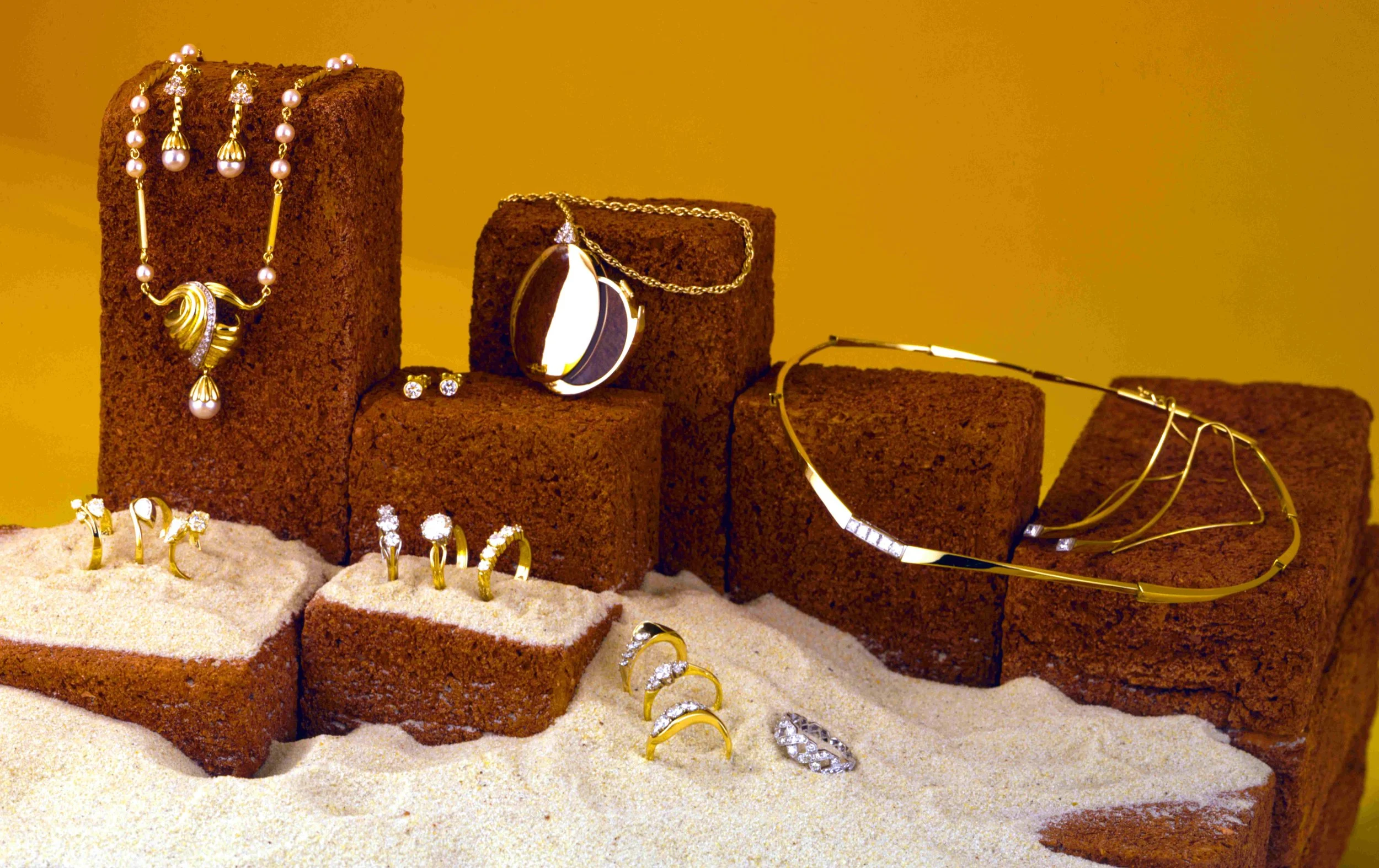 Gold and silver jewelry, including necklaces, rings, earrings, and bracelets, displayed on brown brick and sandy surfaces against a yellow background.