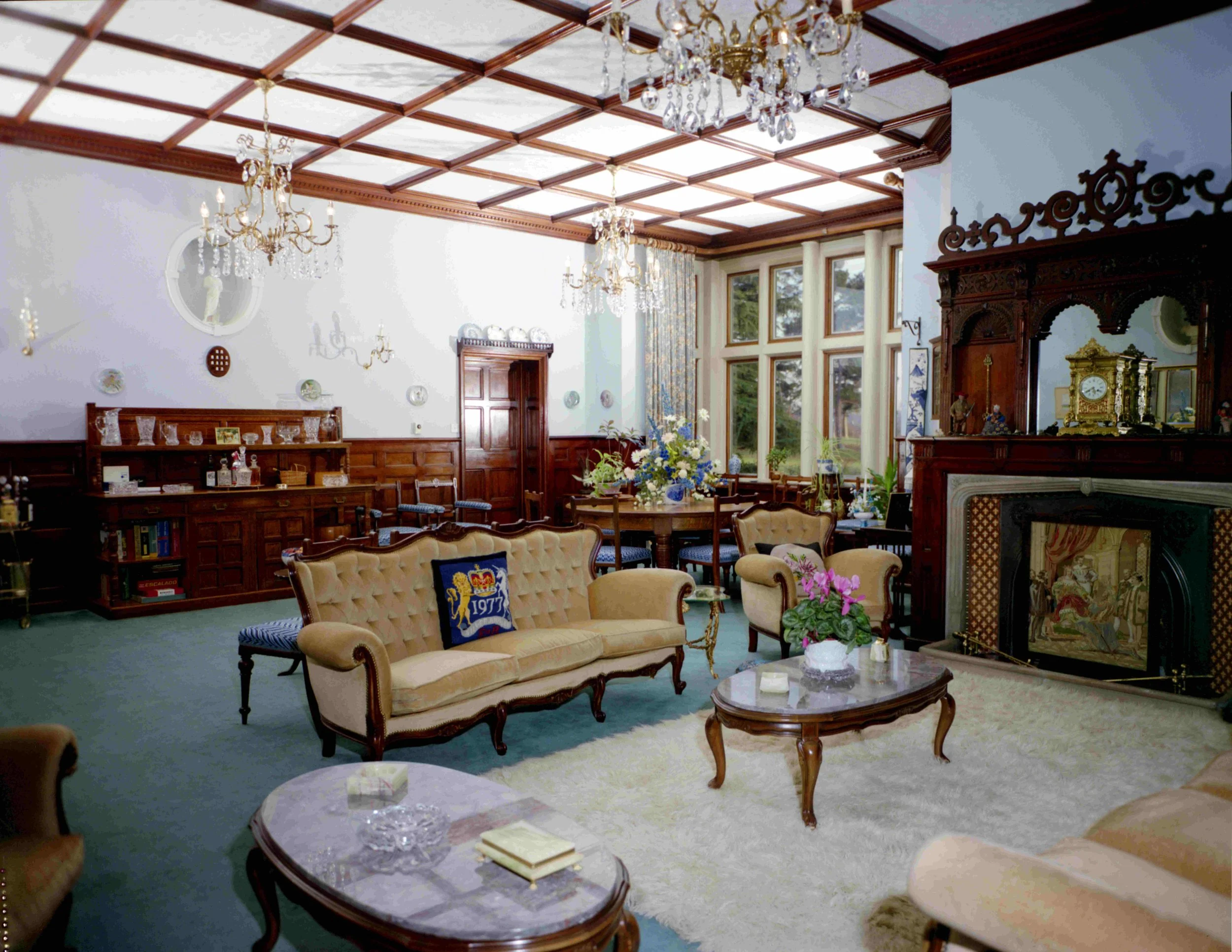 A high-ceiling room with large windows, ornate woodwork, and elegant chandeliers. The room is decorated with vintage furniture, including a beige tufted sofa, wooden tables, and a decorated fireplace. There are floral arrangements and decorative plat
