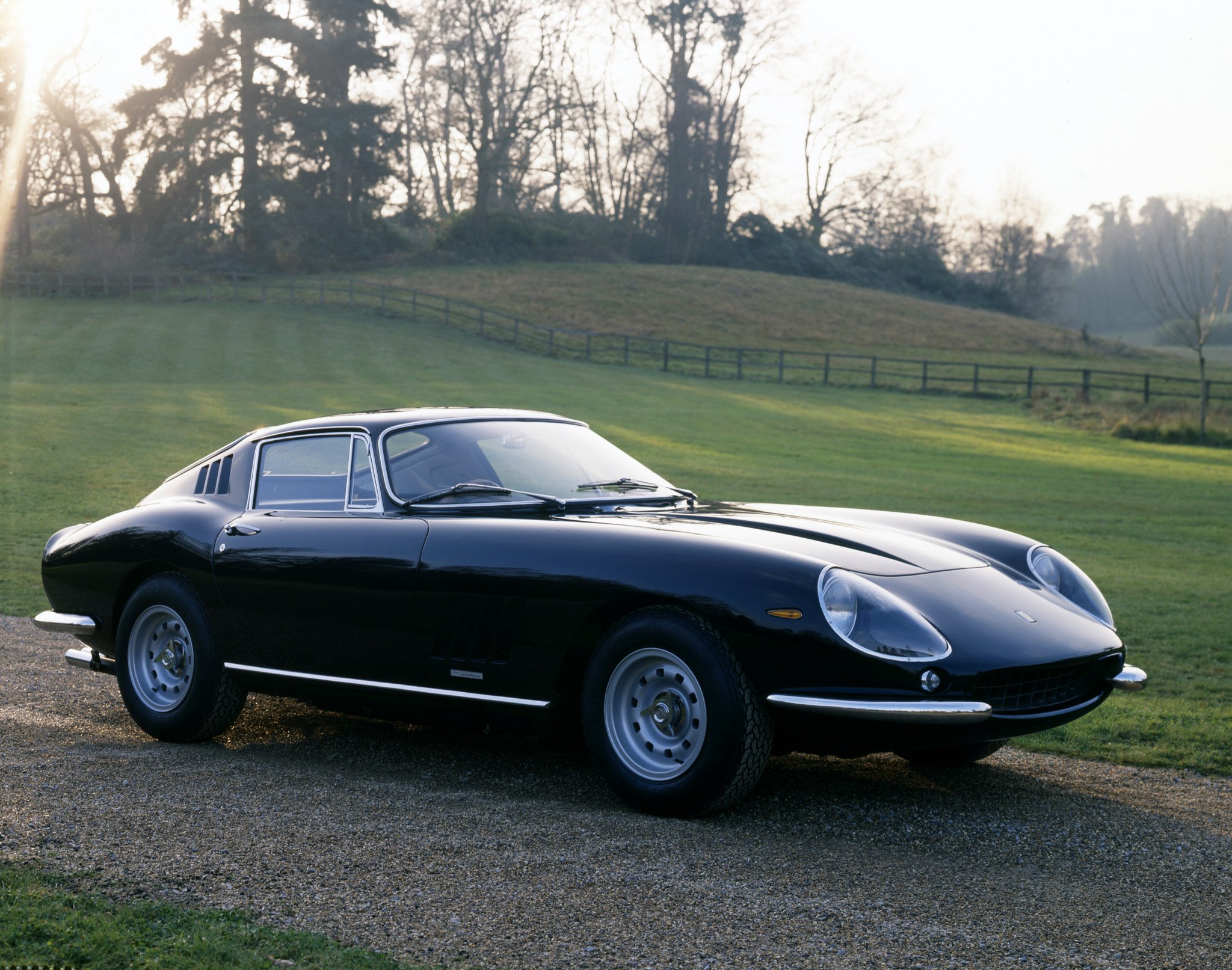 A black vintage sports car parked on a grassy area with a wooden fence in the background and trees in the distance under a bright sky.