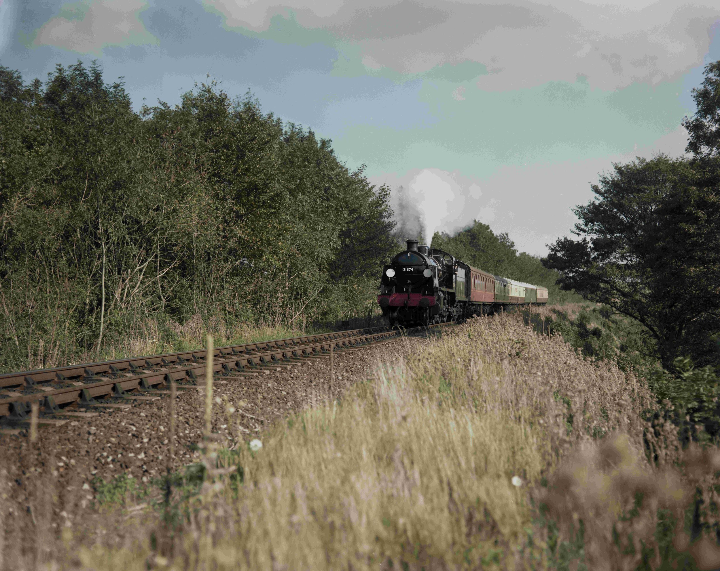 A vintage steam train traveling along a railway track through a green, wooded countryside with a cloudy sky overhead.