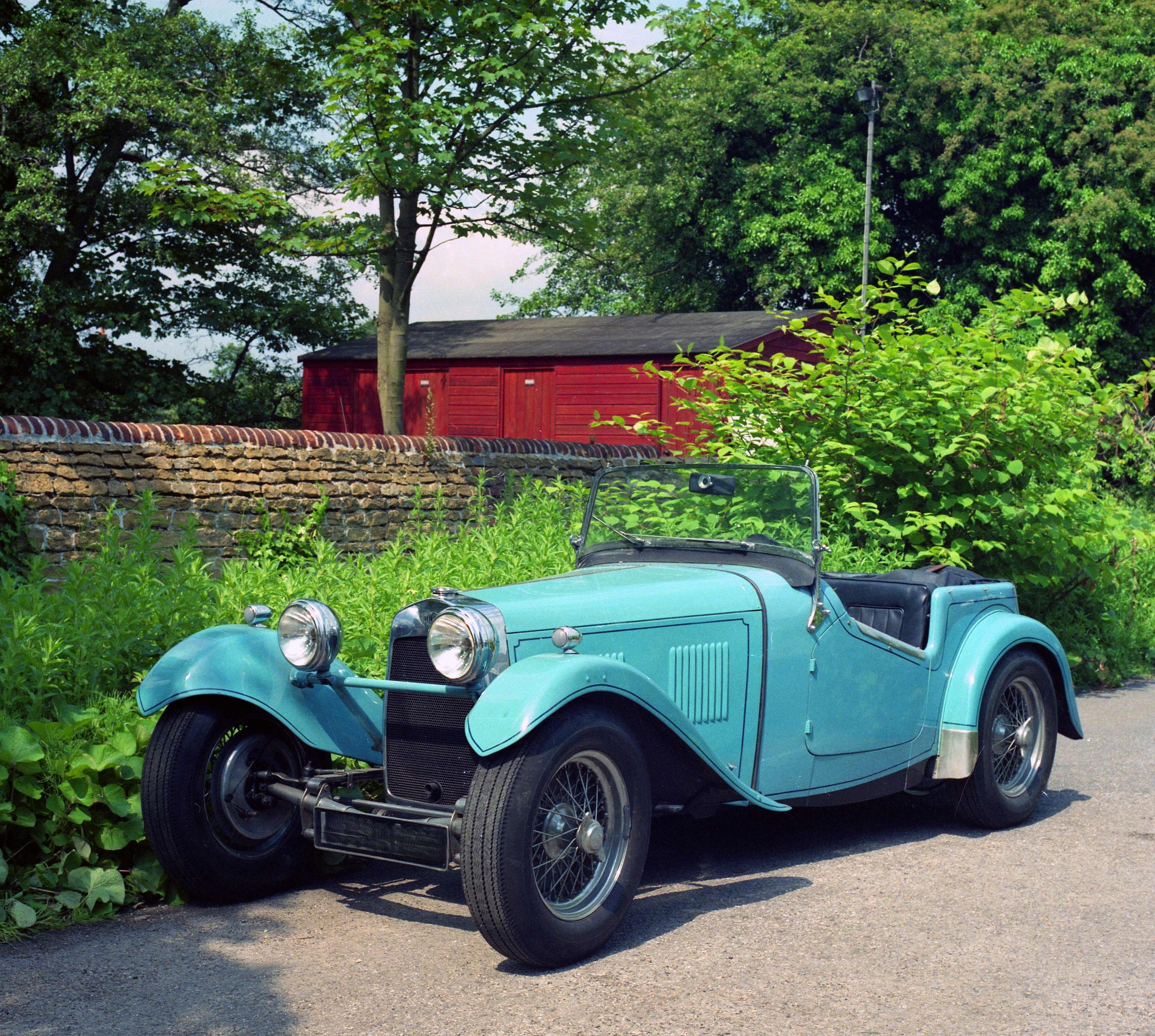 A vintage light blue convertible car parked on a paved street, with a background of dense green trees, a red building with a shingled roof, and a stone wall. The car has round headlights, black tires, and wire-spoked wheels.