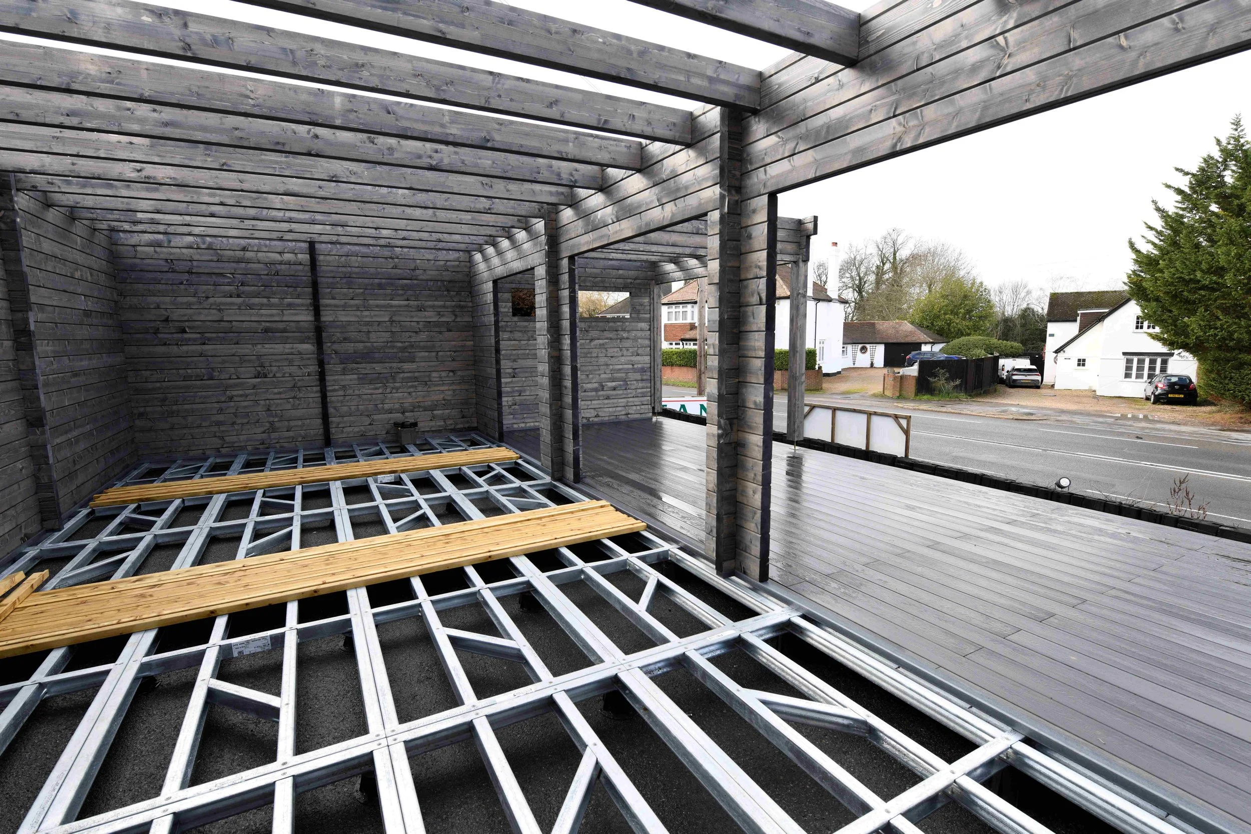 The photo shows the frame of a partially constructed building with wooden beams and metal supports, with an outdoor street scene in the background including houses, trees, and parked cars.