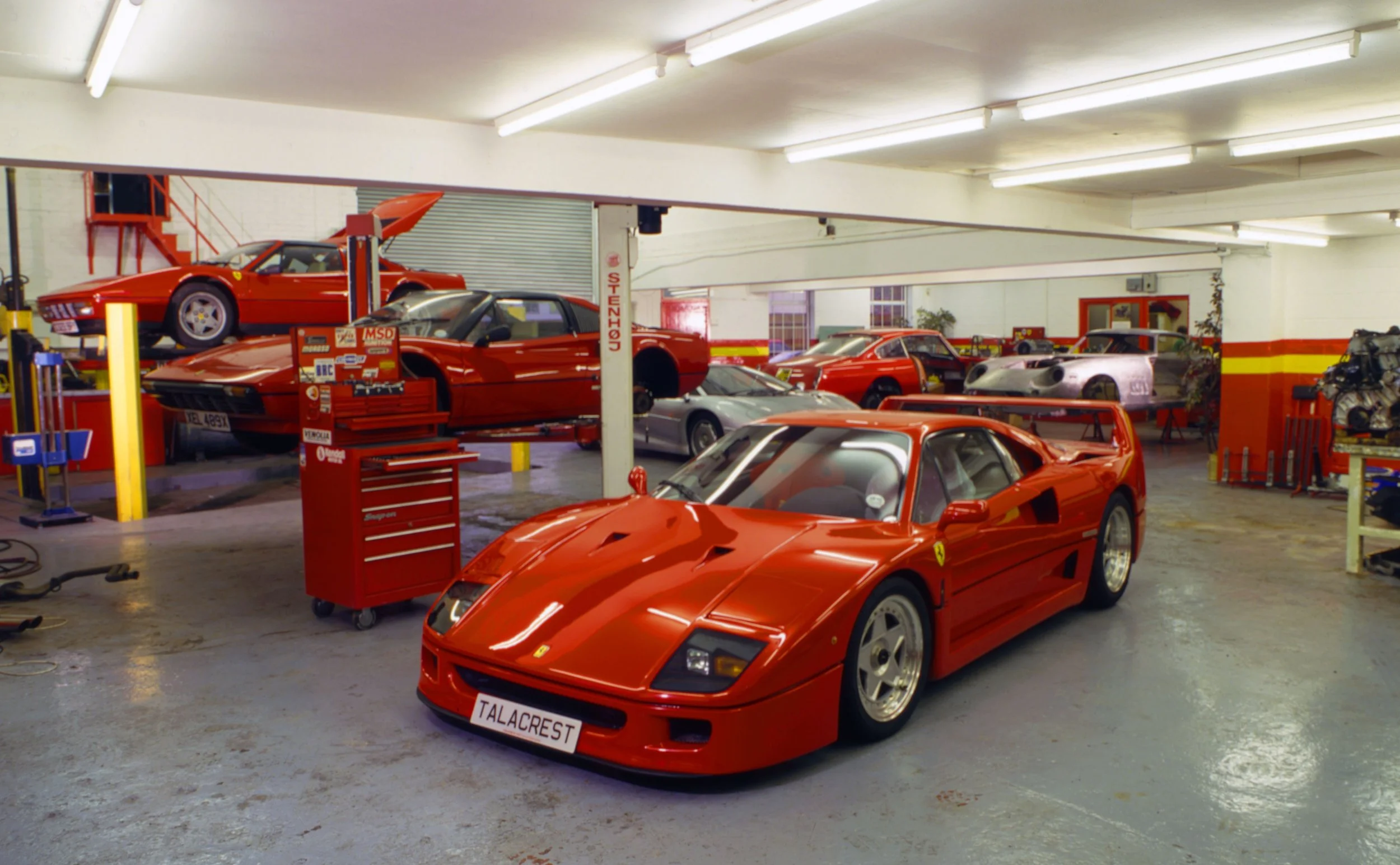 A garage workshop filled with red Ferrari sports cars, some on lifts and others parked on the floor, with tools and car parts visible around.